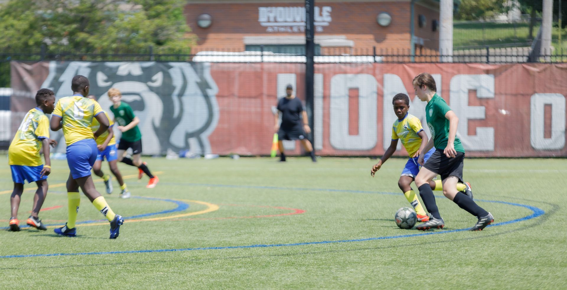 Soccer players in green and blue jerseys compete on a field near a mural-covered wall