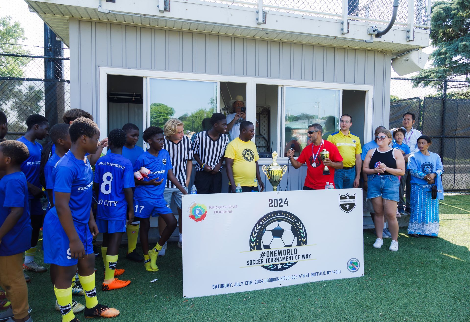 Soccer teams pose with a trophy and banner outside a building on a turf field