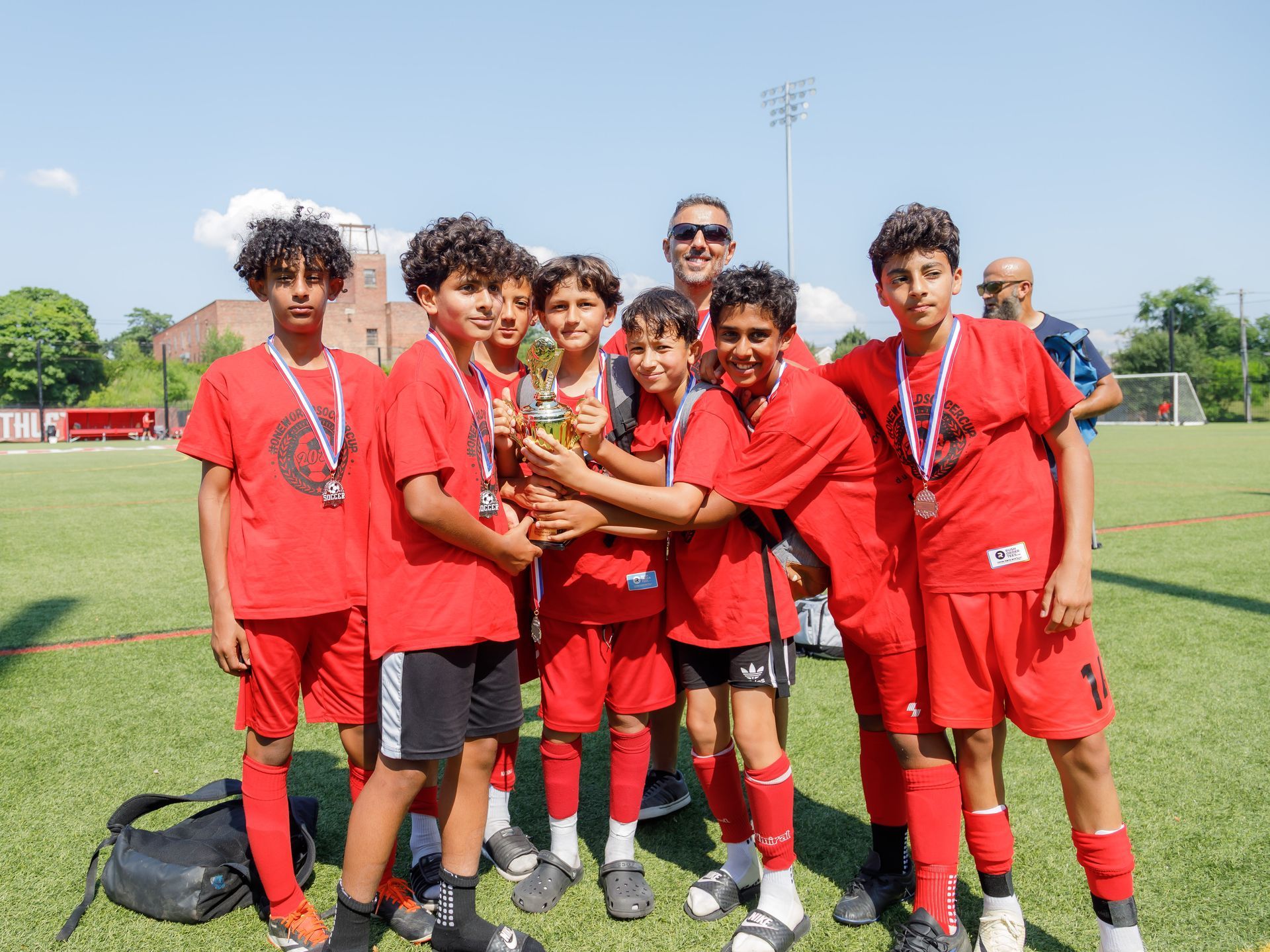 Youth soccer team in red uniforms celebrating a trophy on a field with a coach in the background