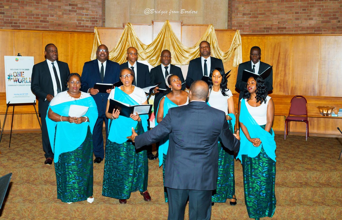 Choir group singing on a church stage, led by a conductor in a suit, with gold drapery backdrop.