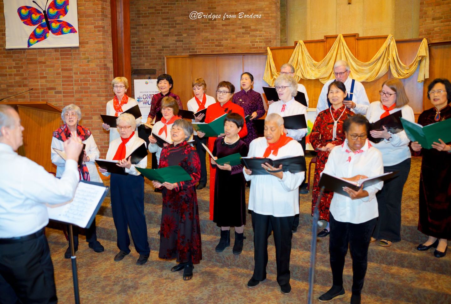 Choir singing with music folders in a brick-walled hall, led by a conductor in front of them.
