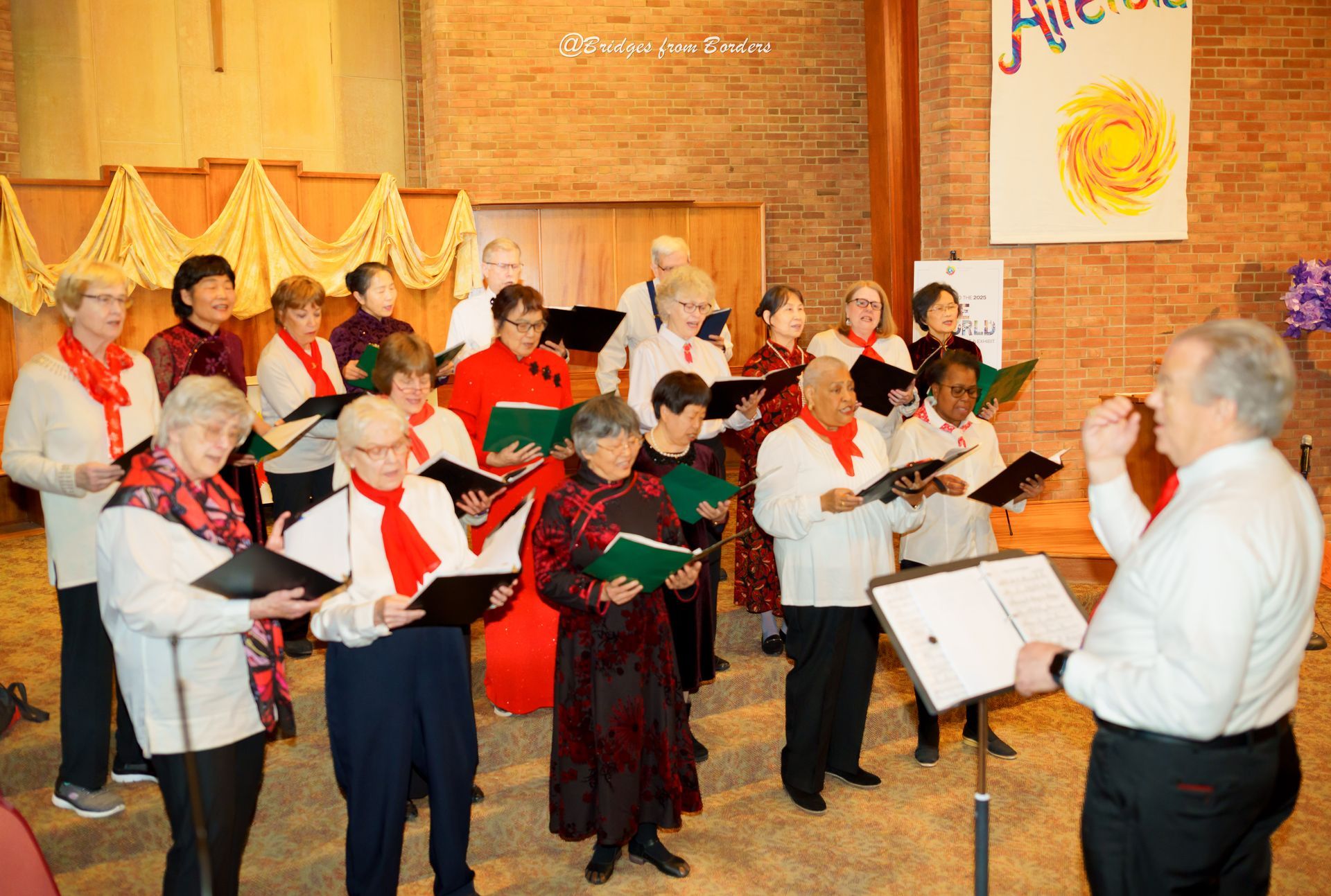 Choir singing from songbooks in a warmly lit hall, conductor at front right, festive decorations on brick wall.
