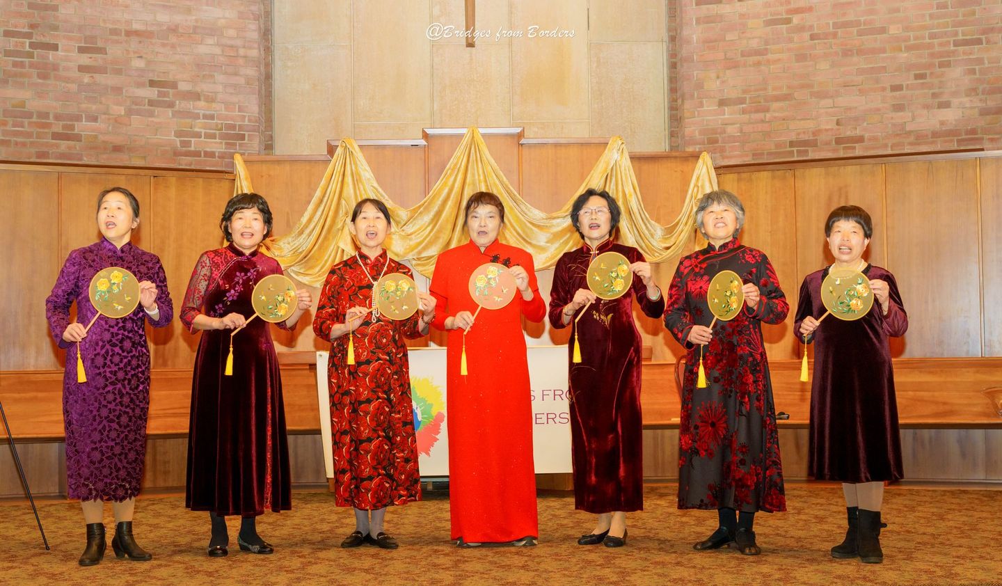Seven performers in colorful traditional dress stand holding fans on a stage with gold drapes.