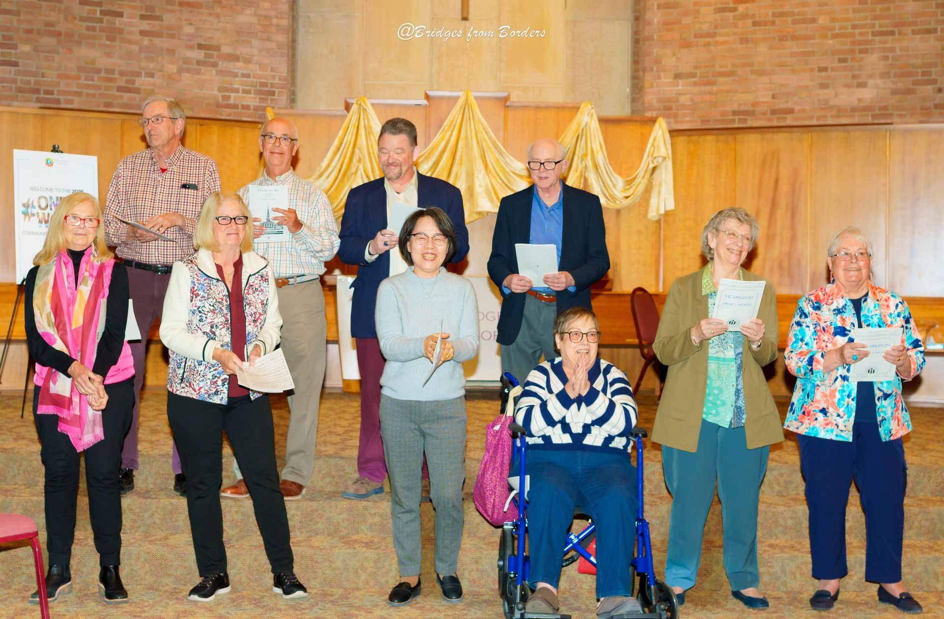 Group of people standing indoors on a stage, holding certificates and gift bags, in front of a wooden wall.