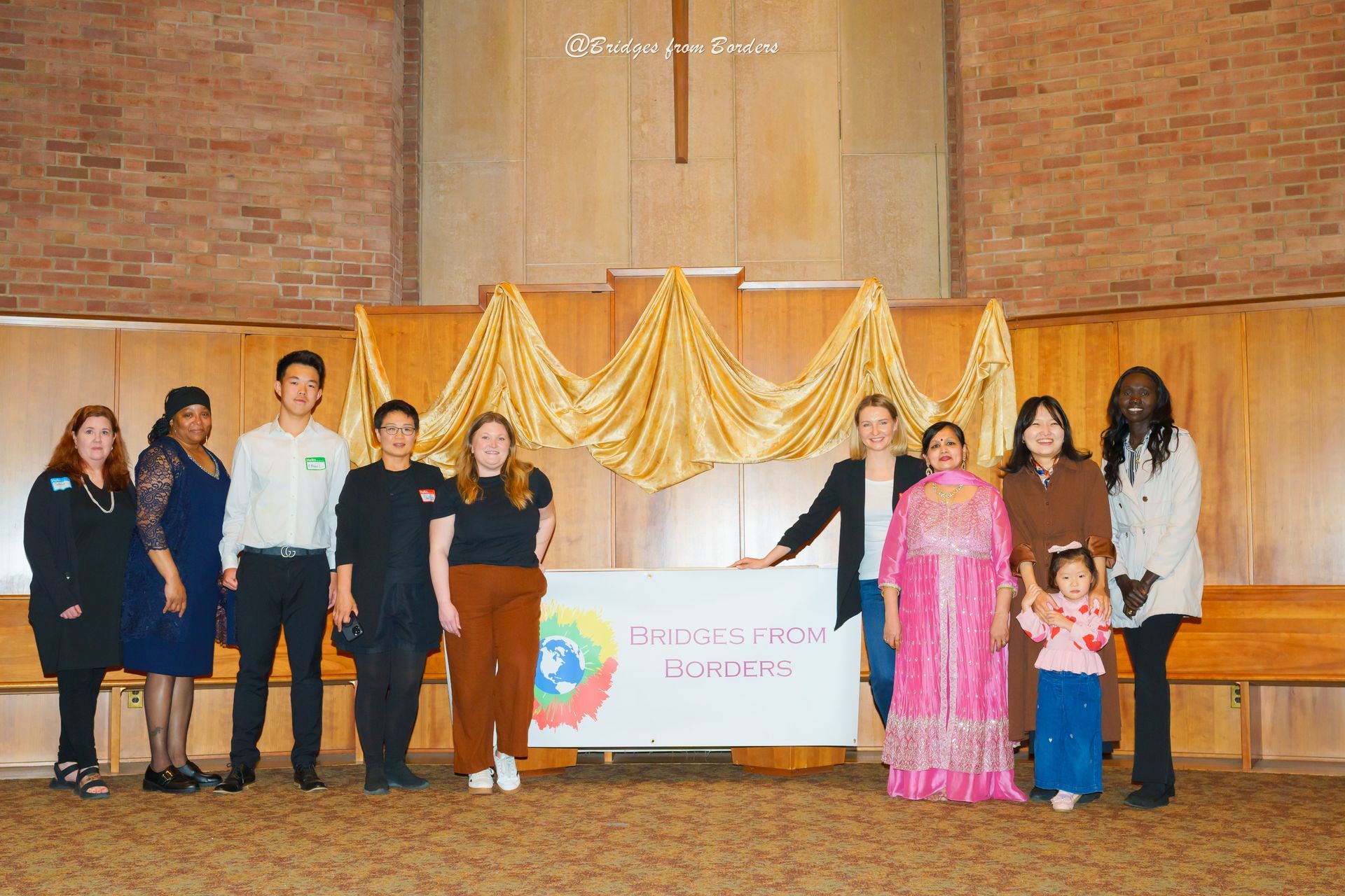 People standing on a stage in front of a gold draped backdrop and banner at an indoor event