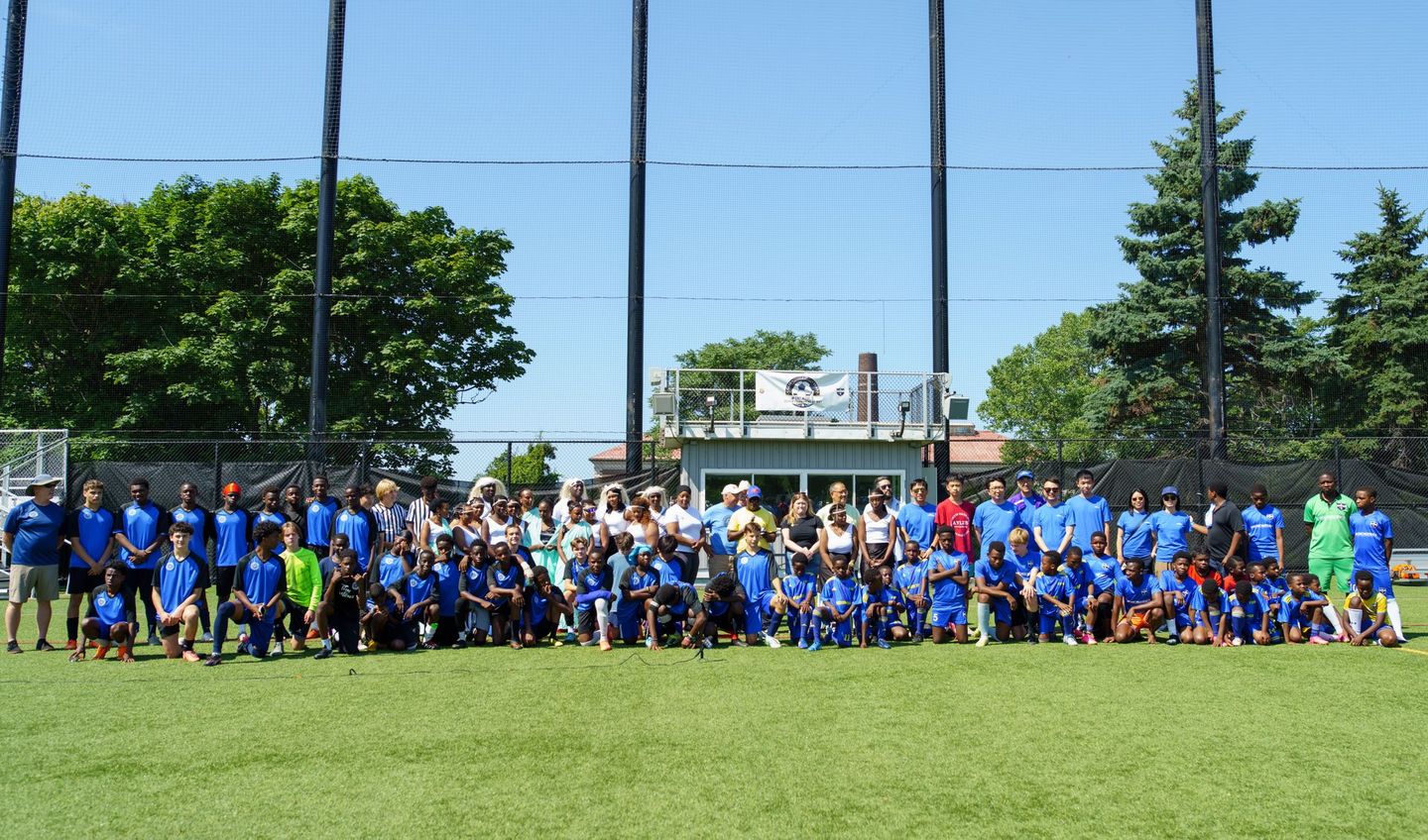 Large group posing on a soccer field in blue shirts under a clear sky, with a fence and trees behind them
