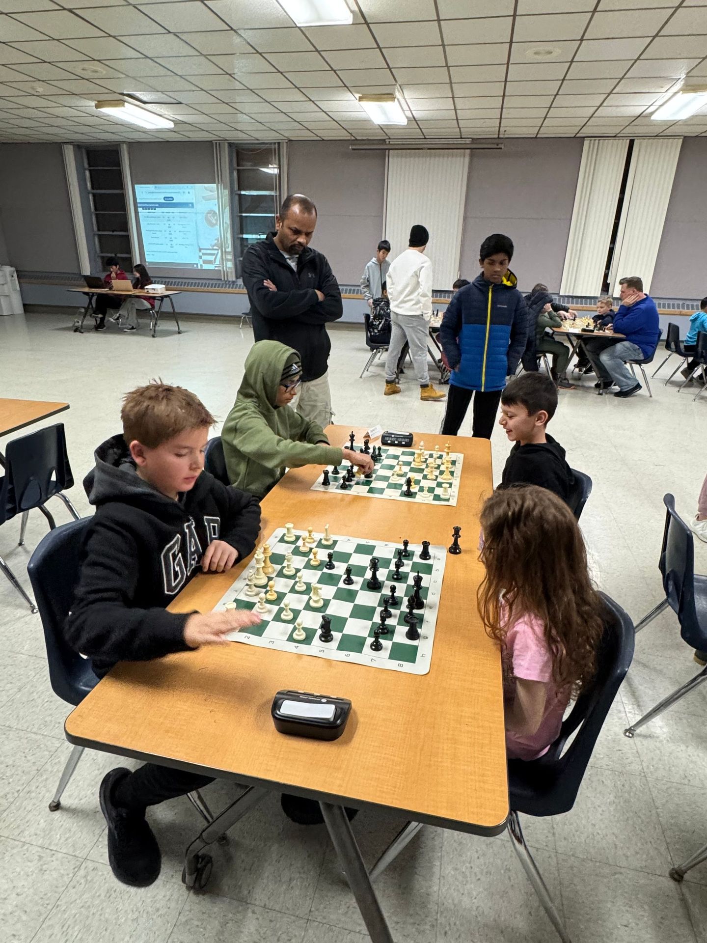 Students play chess at tables in a school cafeteria while adults watch in the background.