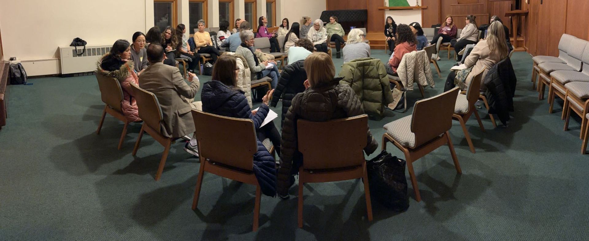 People seated in a circle in a meeting room, viewed from the back, with a speaker at the front.