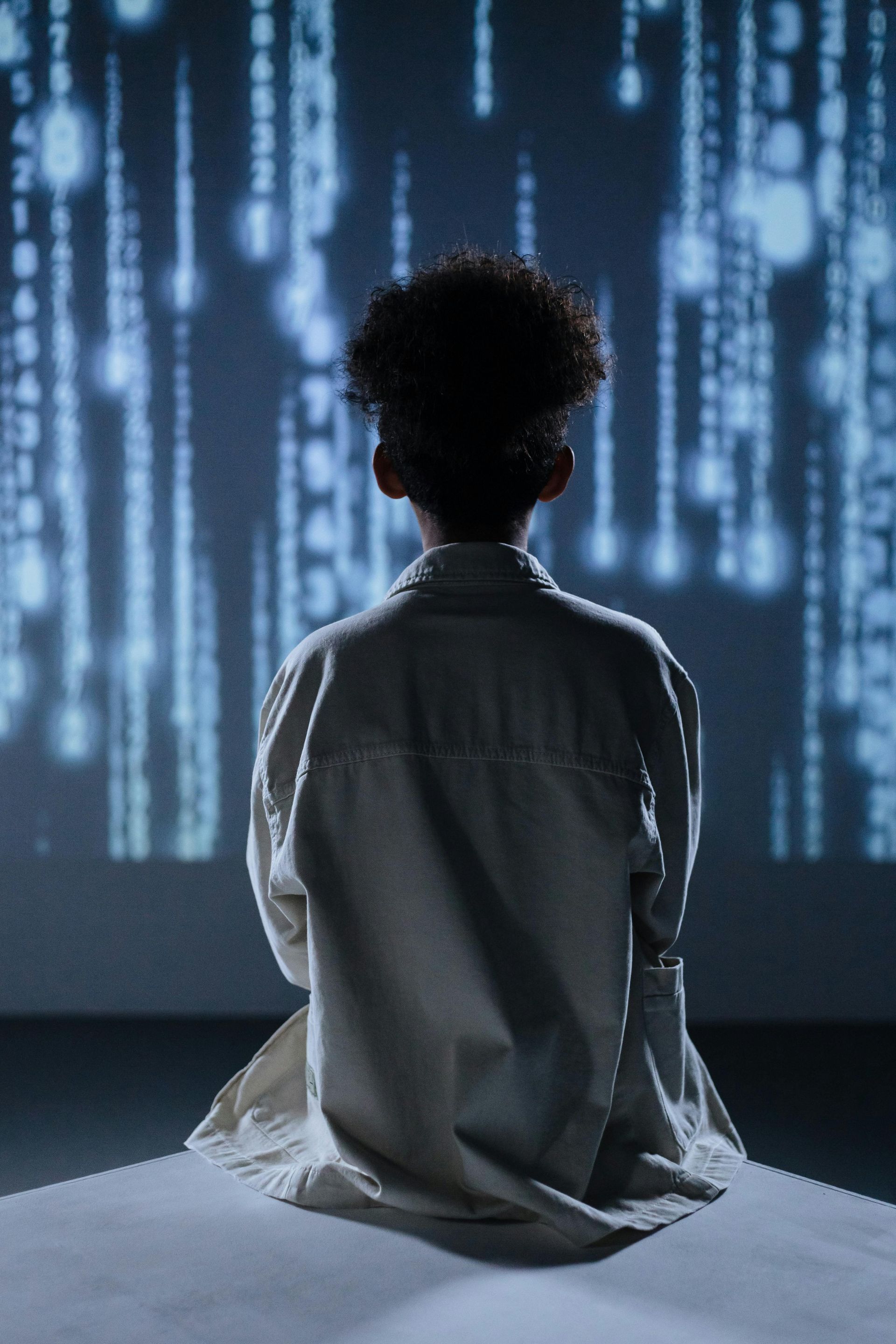 Person seated on a bed, facing glowing blue code on a dark screen