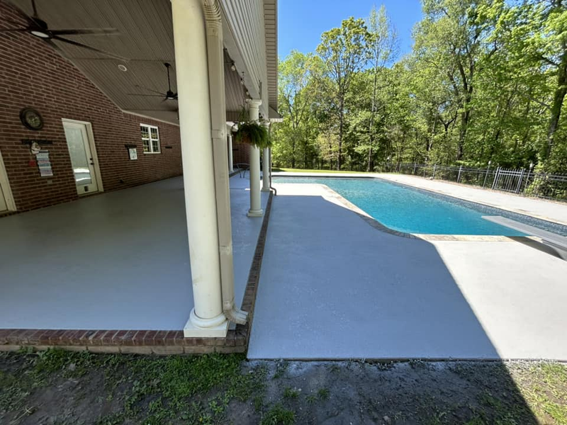 A covered brick patio with white columns overlooks a rectangular blue swimming pool surrounded by a light gray pool deck.