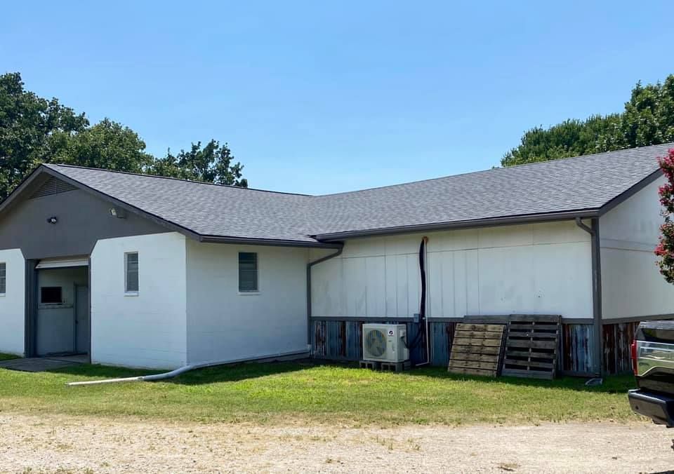 A single-story white building with a gray roof and trim, featuring a side entrance and an exterior AC unit on the lawn.