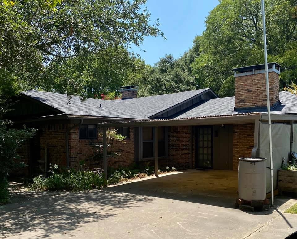 Single-story brick house with a covered patio, grey roof, and chimney, set behind a concrete driveway among trees.