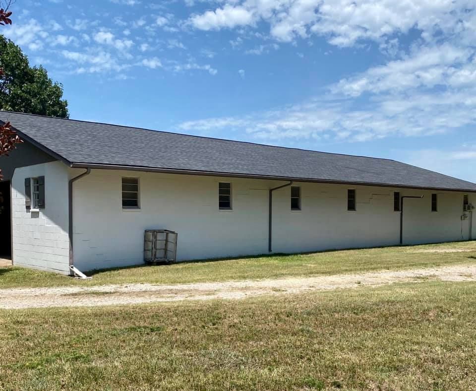 A long, single-story white building with a dark shingled roof, set against a bright blue sky with a gravel path in front.