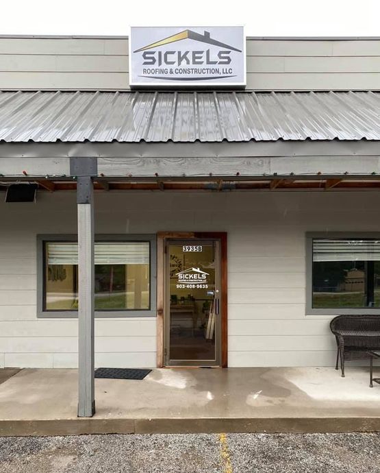 The storefront of Sickels Roofing & Construction, LLC with a grey metal roof and a gravel parking lot in front.