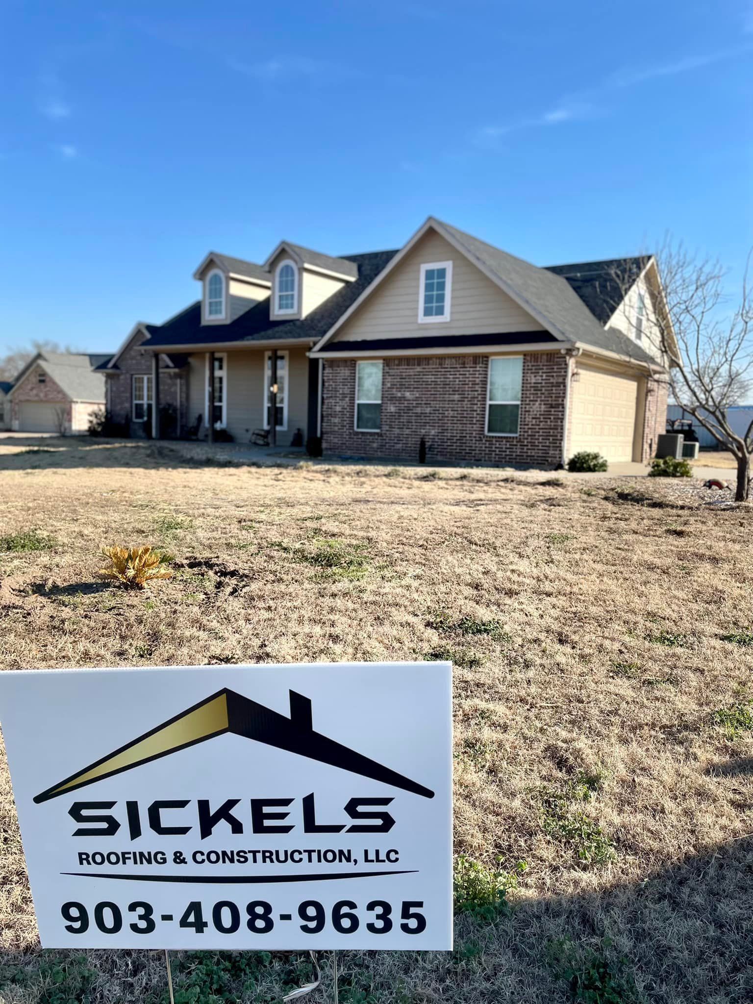 A house with tan siding and brick accents sits on a dry lawn behind a Sickels Roofing & Construction business sign.