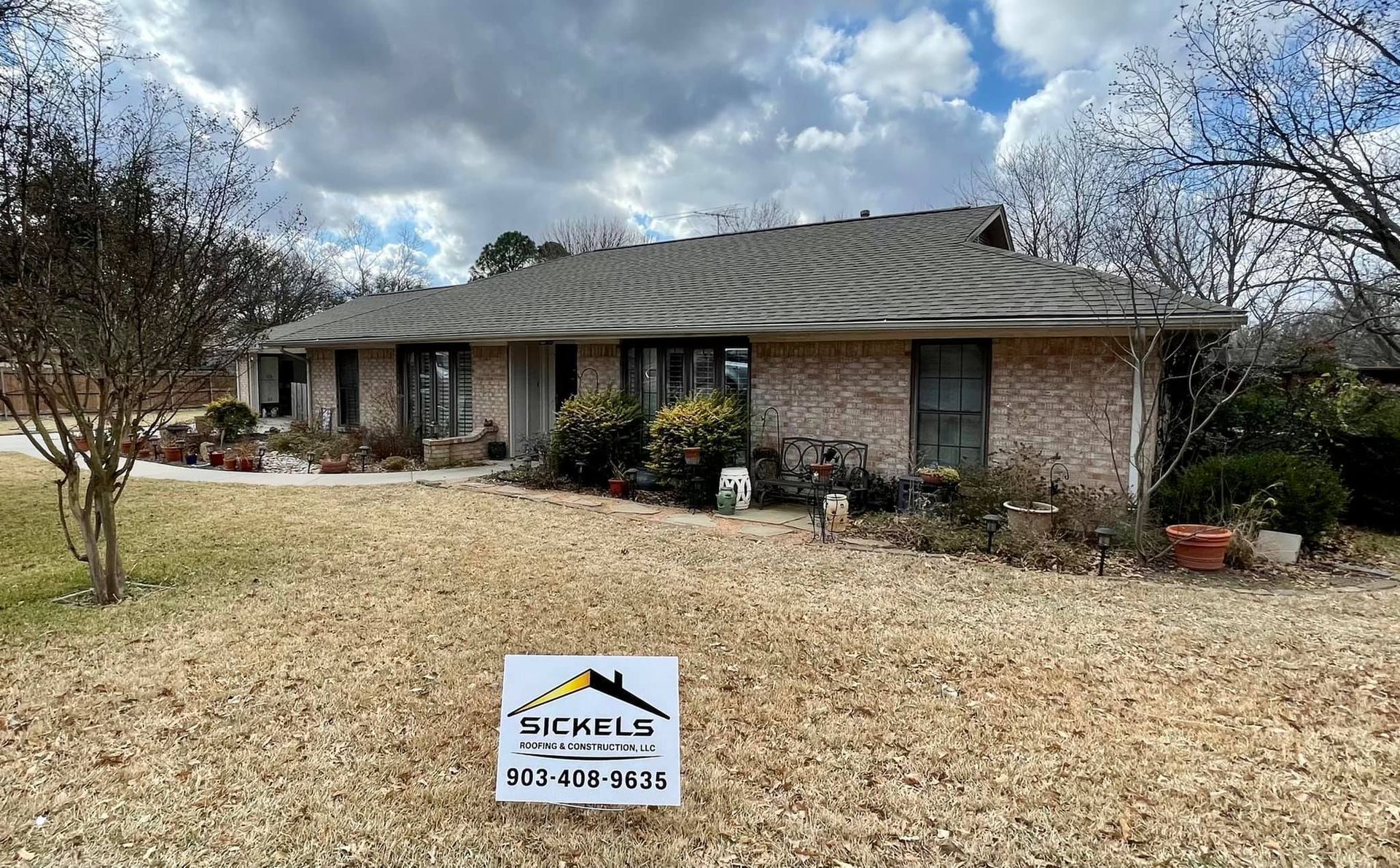 A single-story brick house with a gabled roof and a yard featuring a yard sign for 