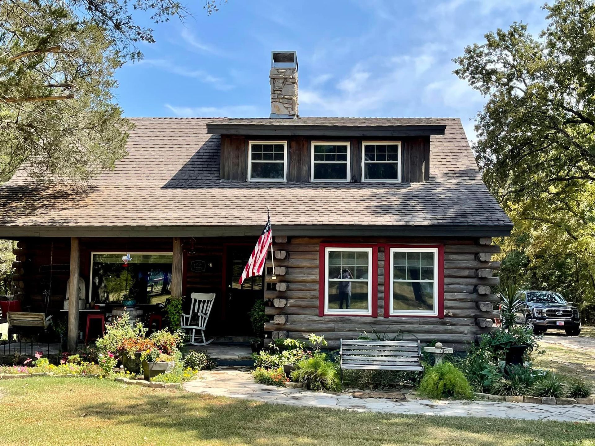 A rustic brown log cabin with a stone chimney, front porch, and dormer windows, surrounded by trees and a stone walkway.