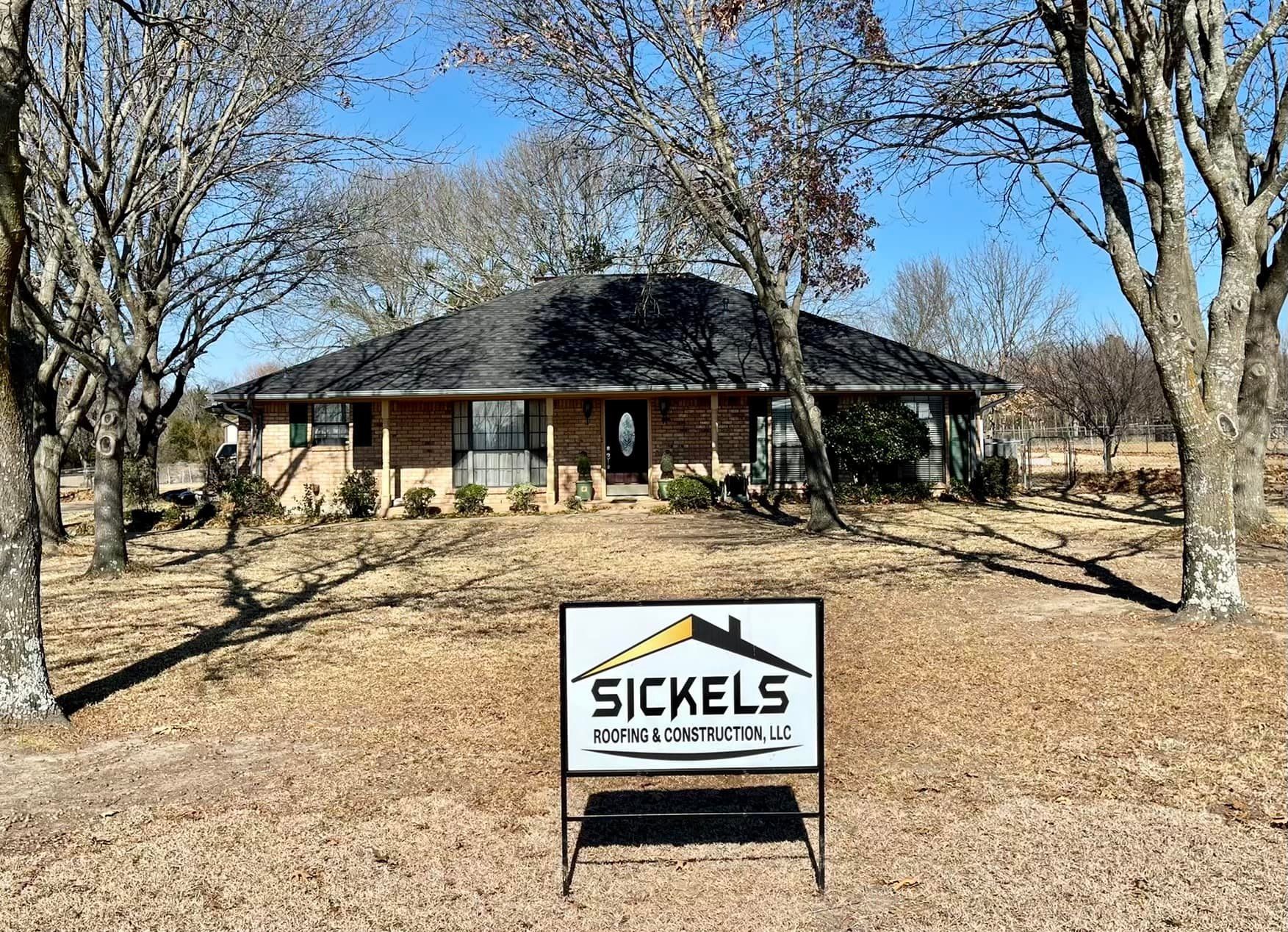 A brick house set back on a dry, grassy lawn under leafless trees, with a Sickels Construction sign in the foreground.