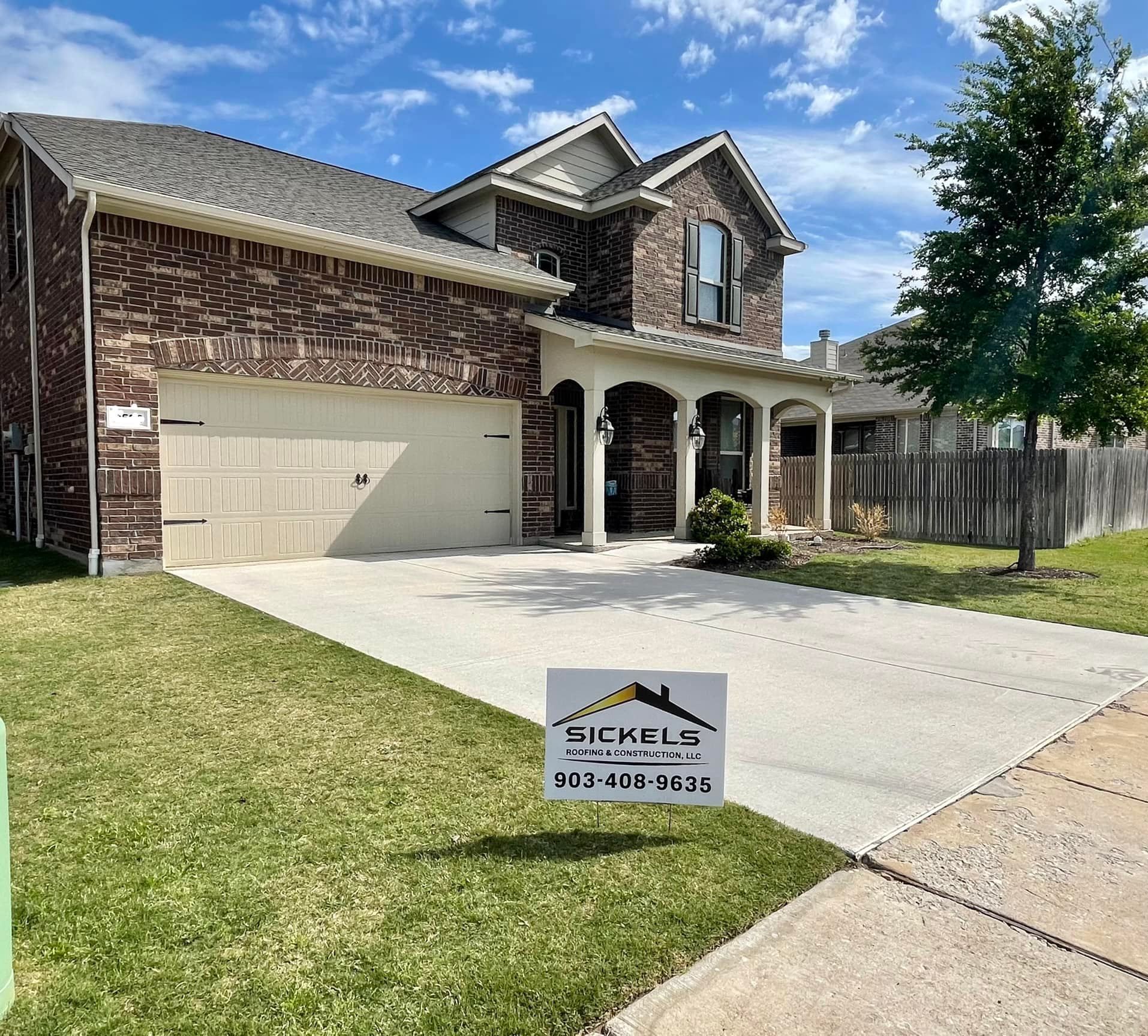 A two-story, brown brick house with a white garage and a lawn sign in a suburban neighborhood under a blue sky.