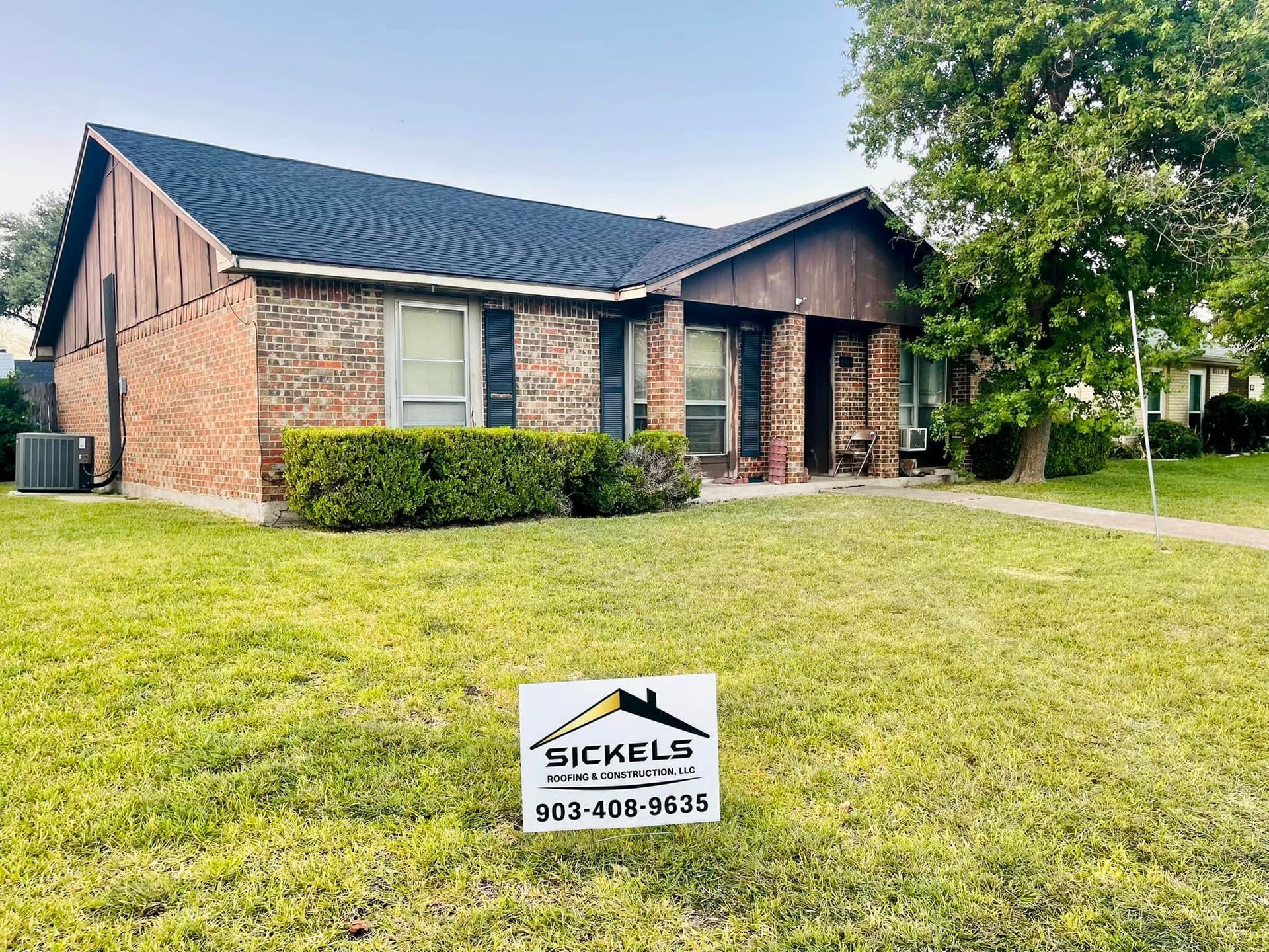 A brick single-story house with a dark roof and a Sickels Roofing yard sign in the front lawn.