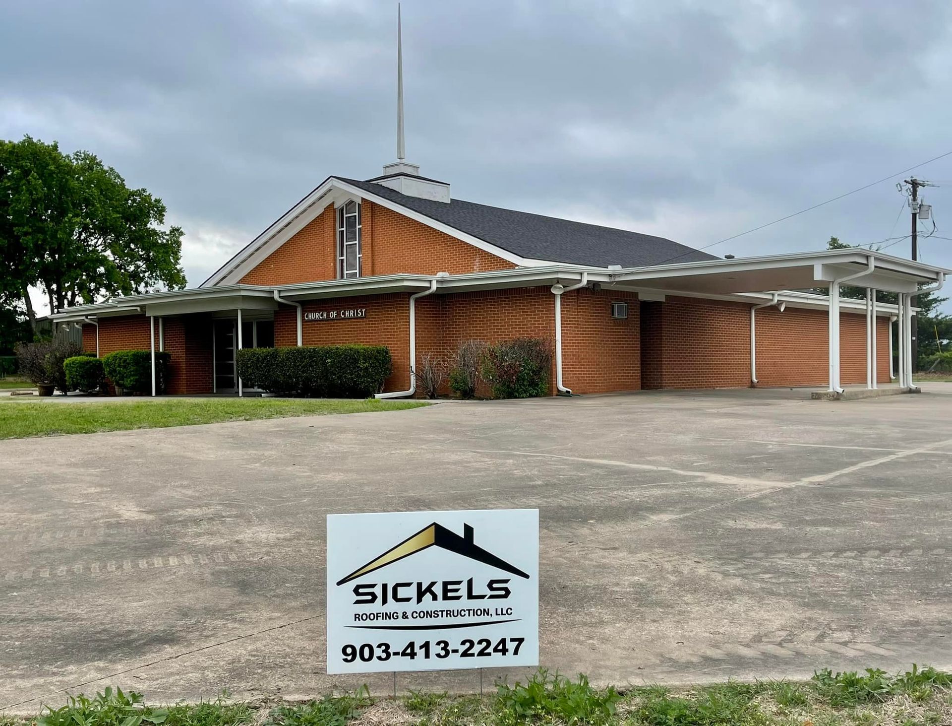 A brick church building with a steeple and covered entrance, featuring a Sickels sign in the foreground parking lot.