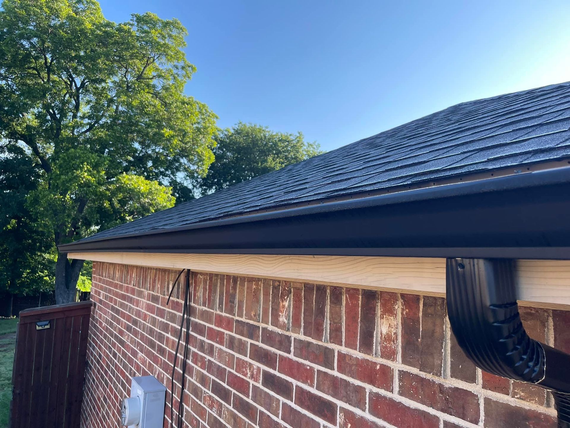 A close-up view of a black gutter system installed on a brick house with a dark shingled roof under a clear blue sky.