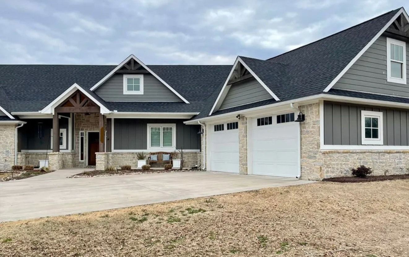 A single-story craftsman home featuring dark gray siding, a stone veneer base, a black shingled roof, and a two-car garage.