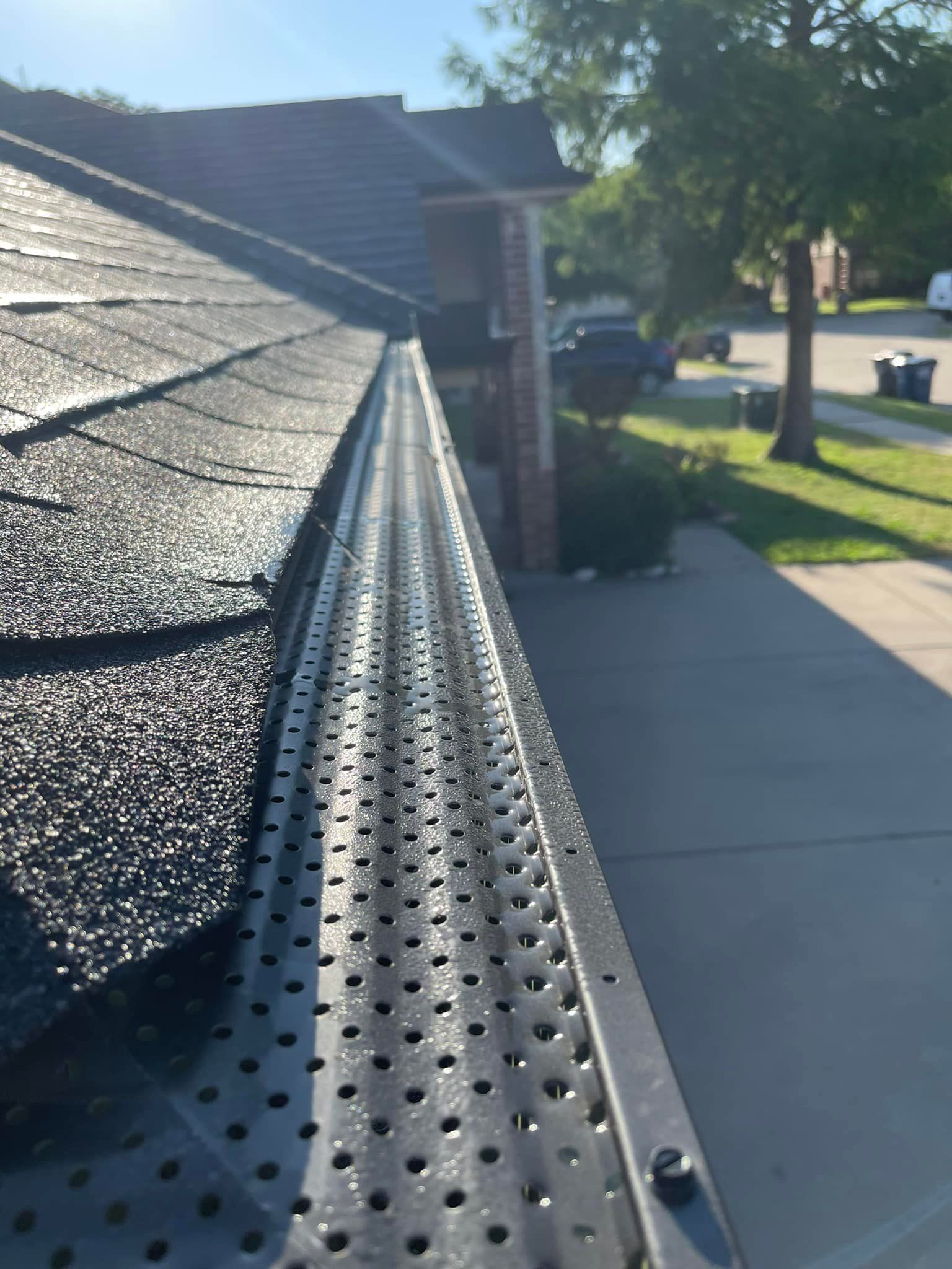 Black metal gutter guard installed over a residential gutter, with a shingled roof and suburban street in the background.