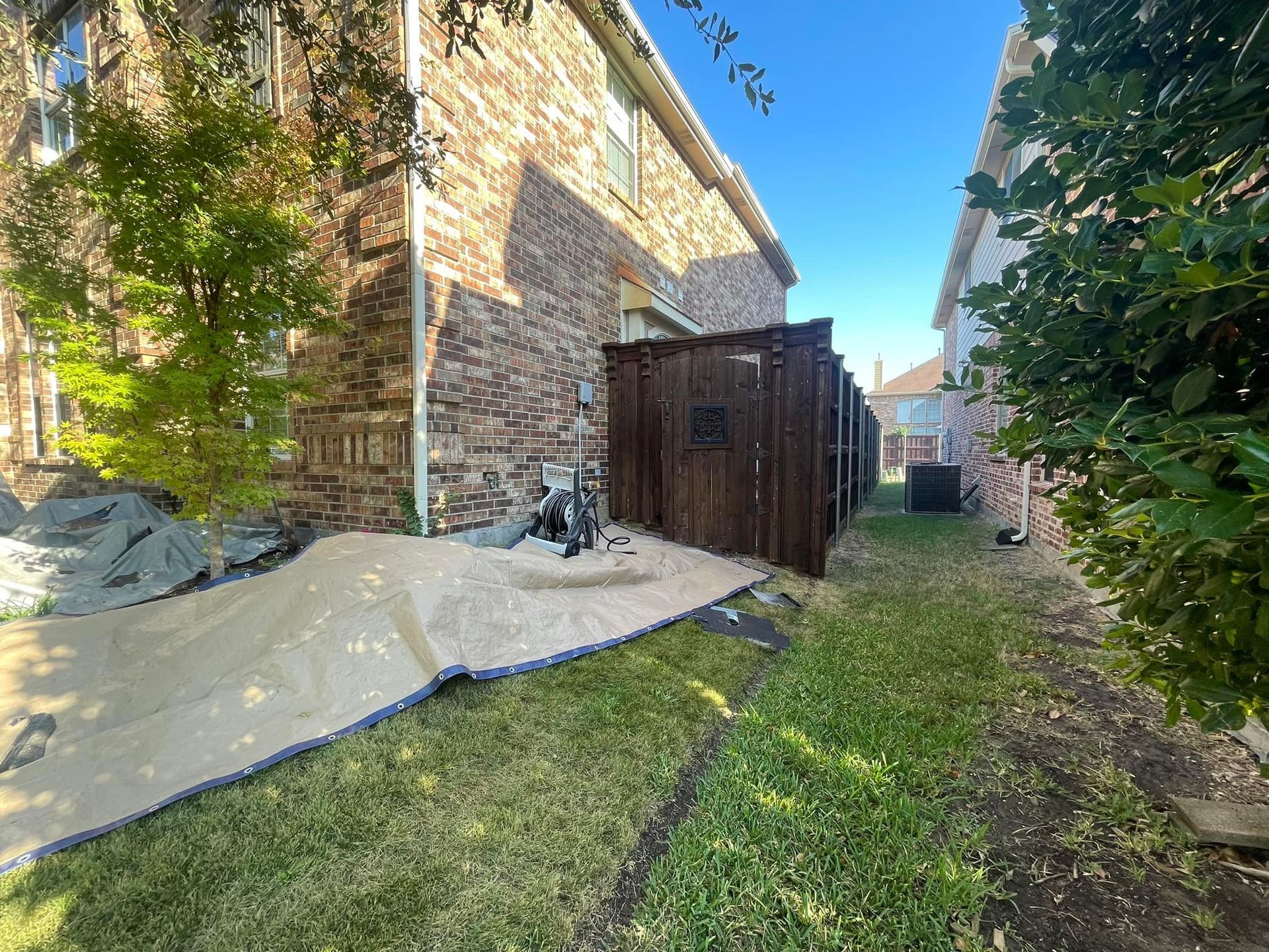 A side yard showing a brick house, a tan landscaping fabric laid on the ground, and a dark wooden fence on a sunny day.