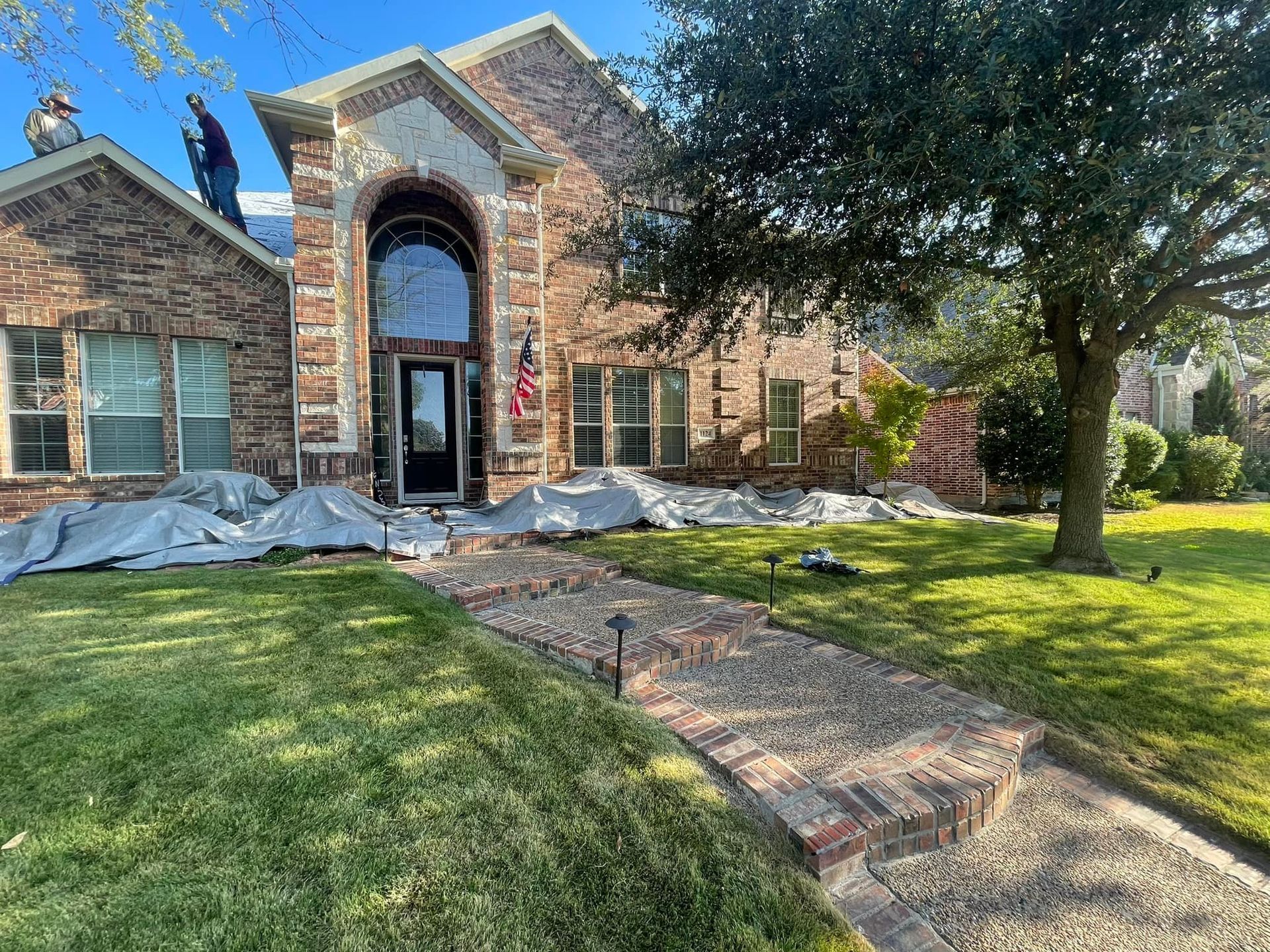 Workers on a brick house roof with protective tarps on the lawn, showing a front entrance and stone pathway.