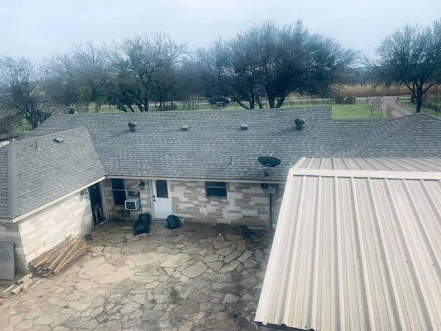 An elevated view of a stone house with a gray shingled roof, a paved patio, and a metal-roofed structure in the foreground.