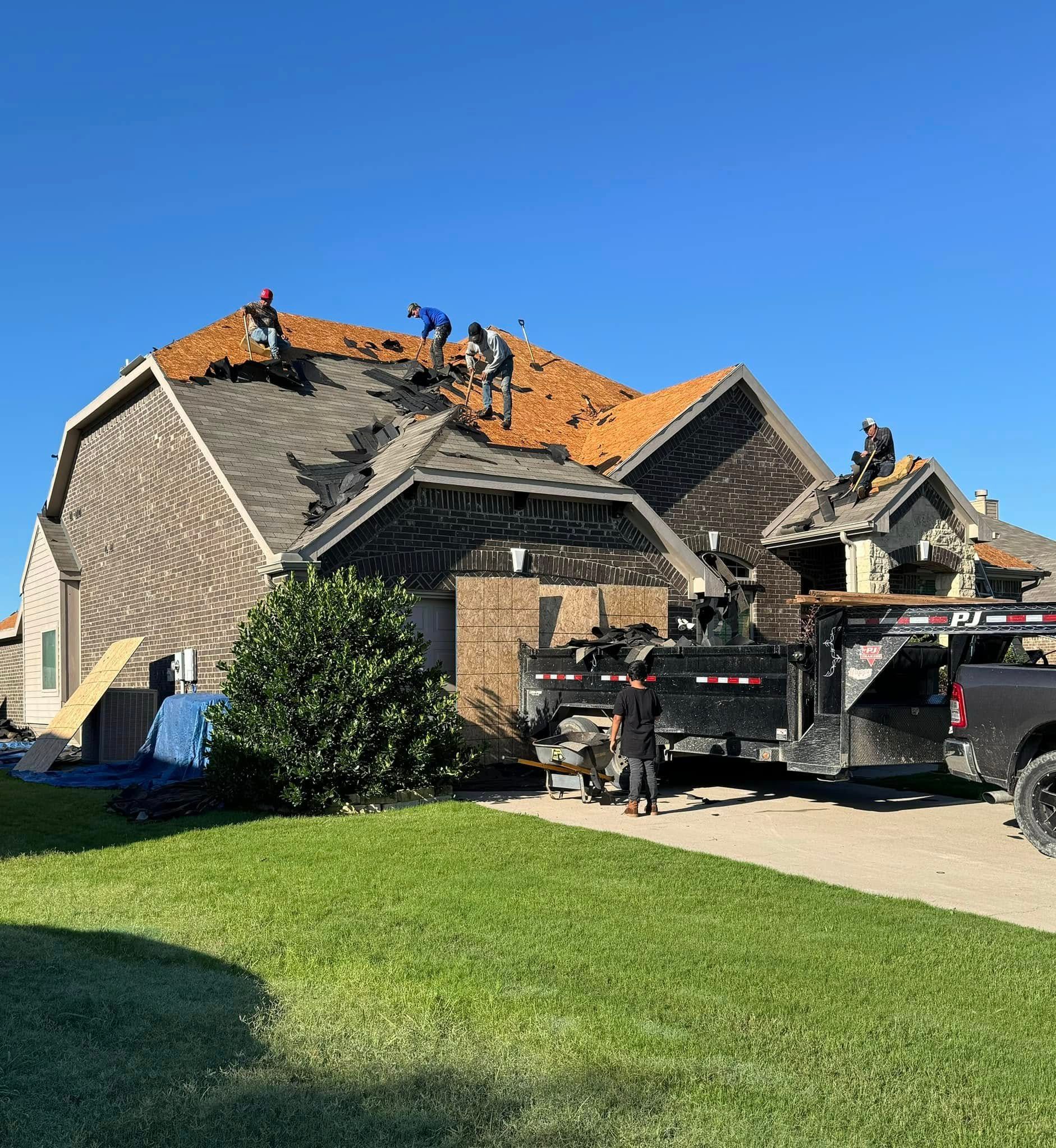 Roofers removing old shingles from a house with a large trailer parked in the driveway on a sunny day.
