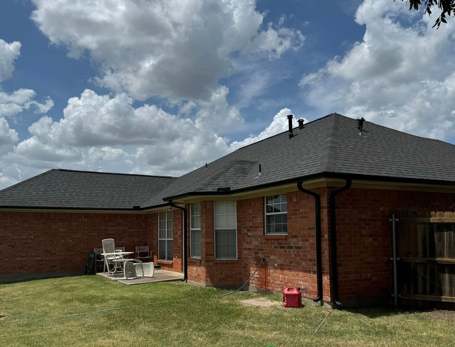 The back of a red brick house with a dark shingled roof, a small patio, and a grassy yard under a cloudy blue sky.