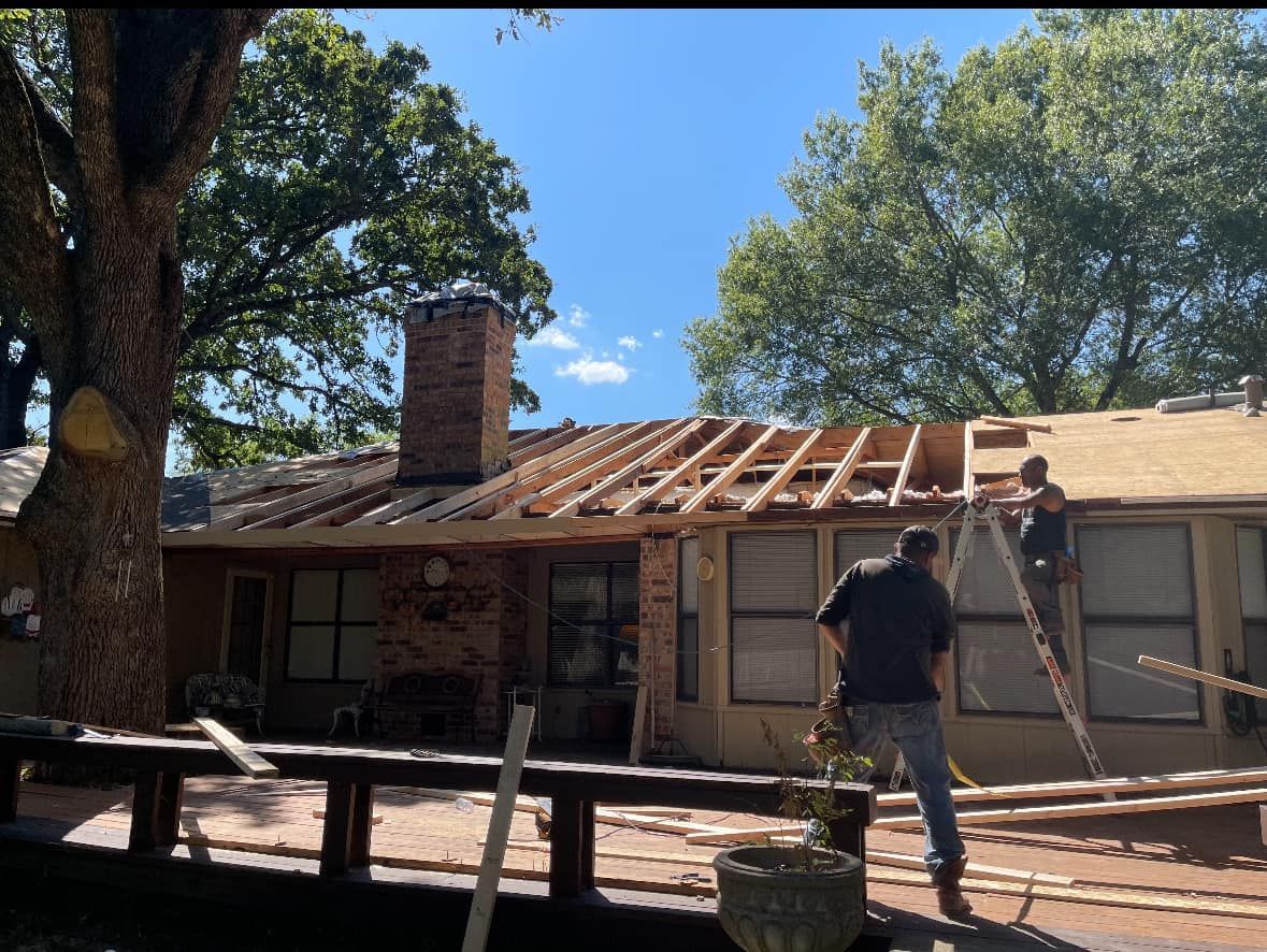 Two workers perform roof repairs on a residential home with a brick chimney under a clear blue sky.