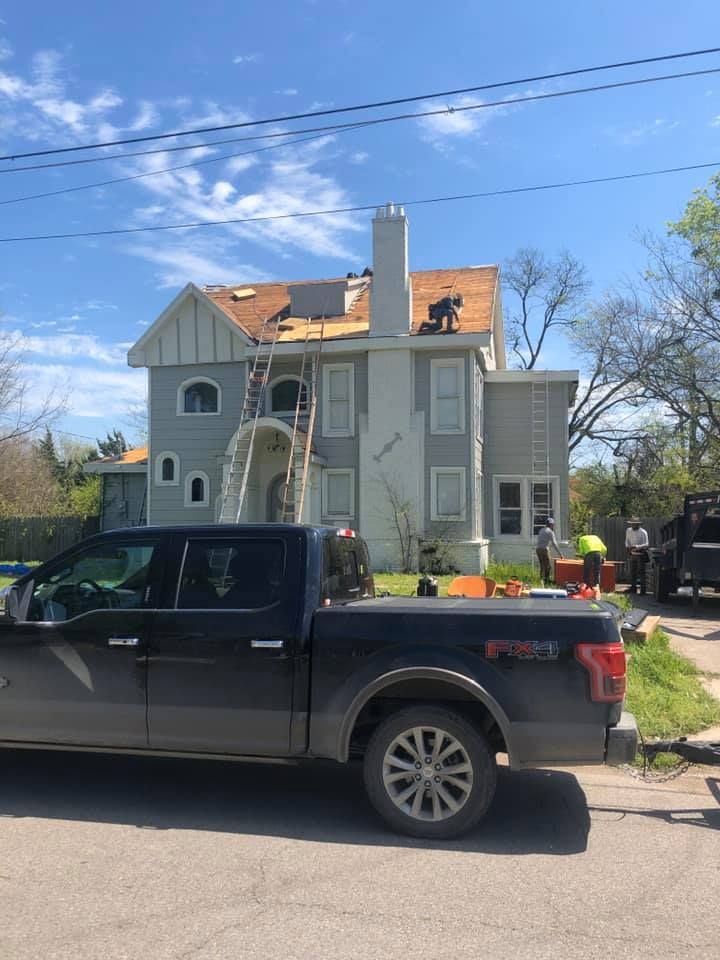 A black pickup truck parked in front of a gray multi-story house undergoing roof repairs with workers on the roof.