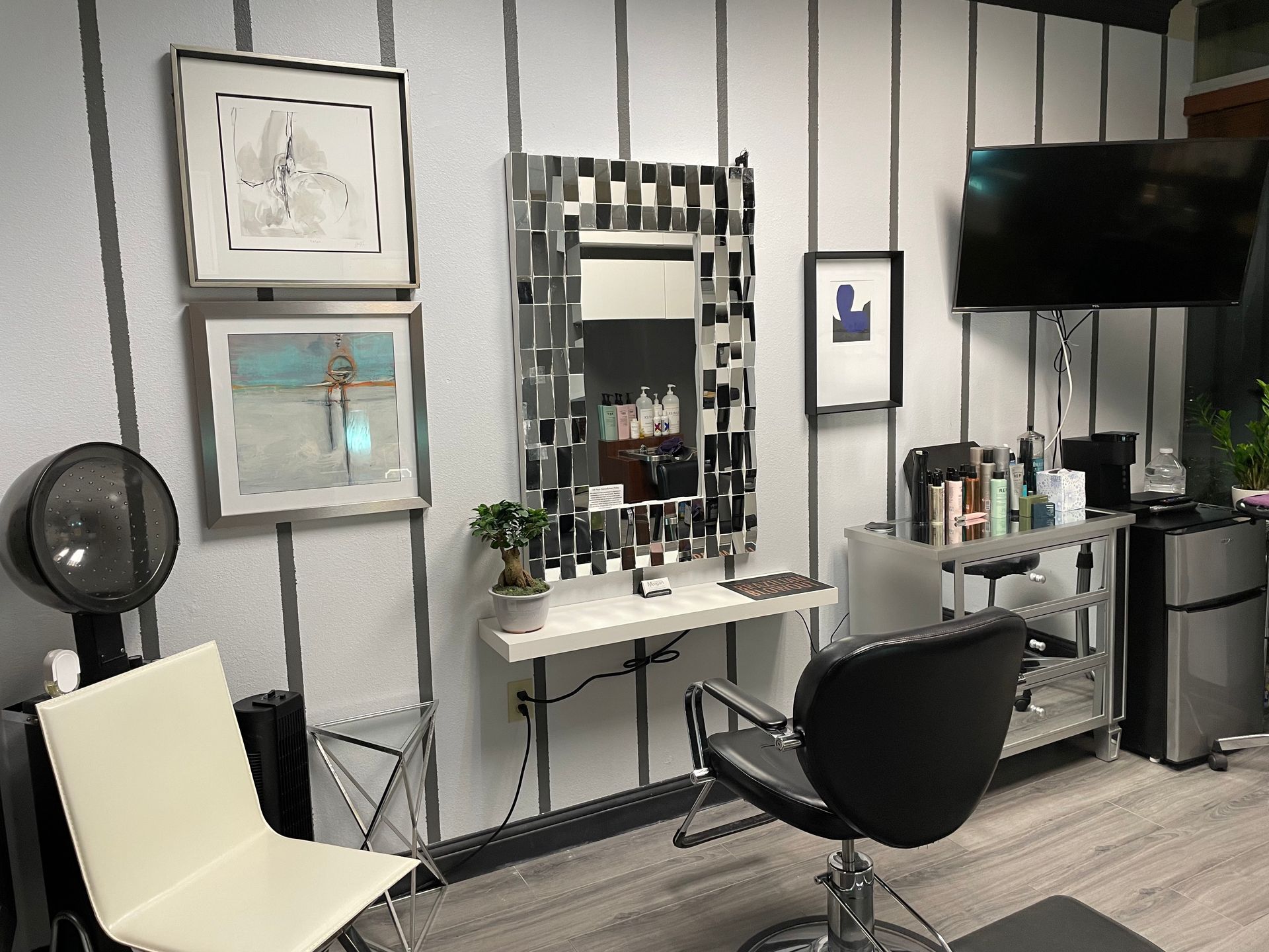 Hair salon interior with mirror, styling chair, artwork, and a small refrigerator.