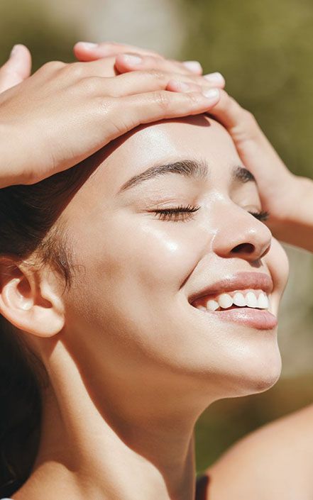 Smiling woman with eyes closed, hands resting on her head in warm sunlight