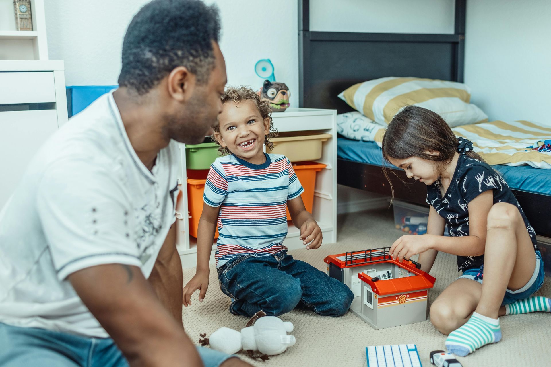 Un homme et deux enfants jouent avec des jouets dans une chambre. Le garçon sourit, la fille joue.