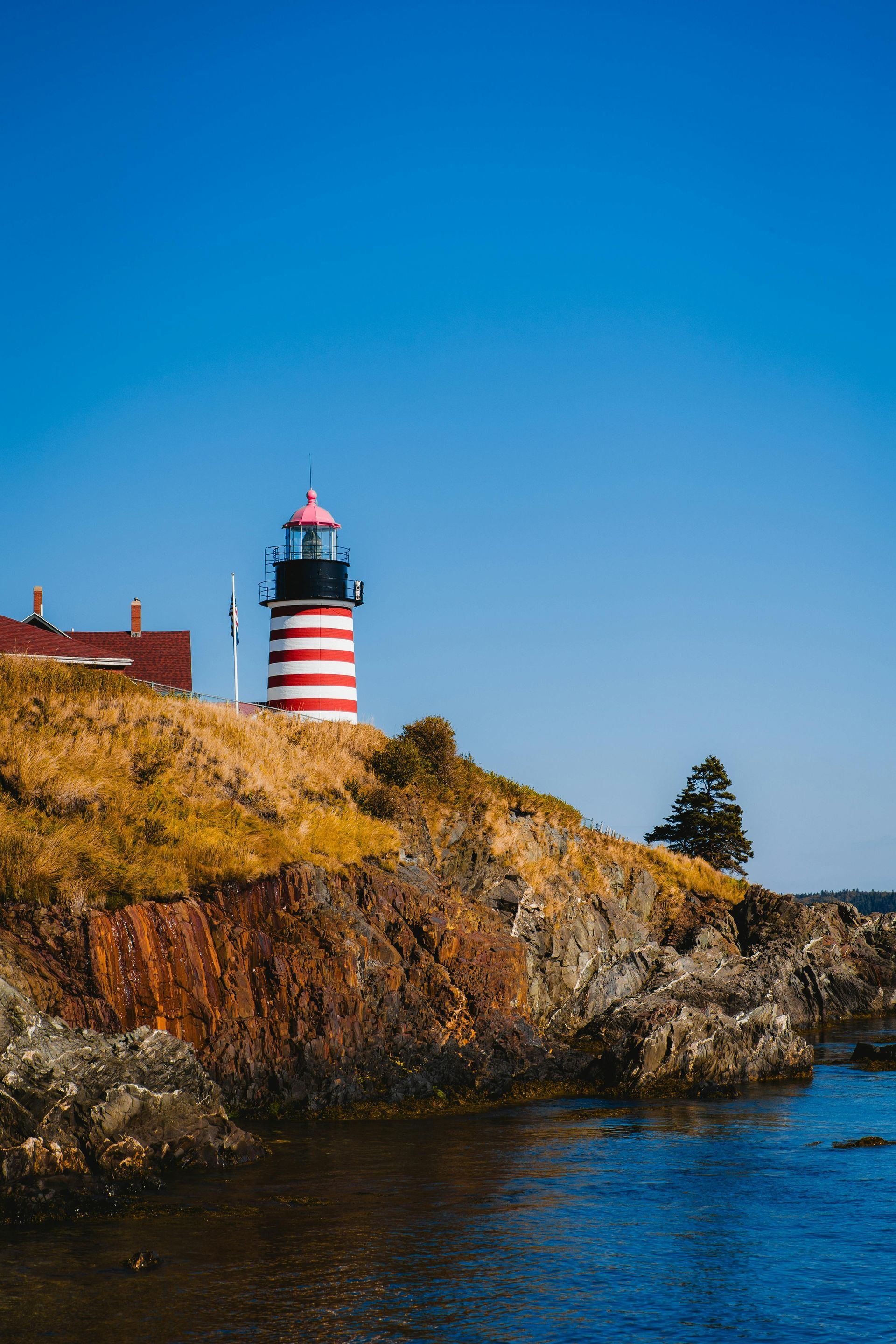 A red-and-white striped lighthouse sits atop a rocky, grass-covered cliff overlooking the ocean under a clear blue sky.