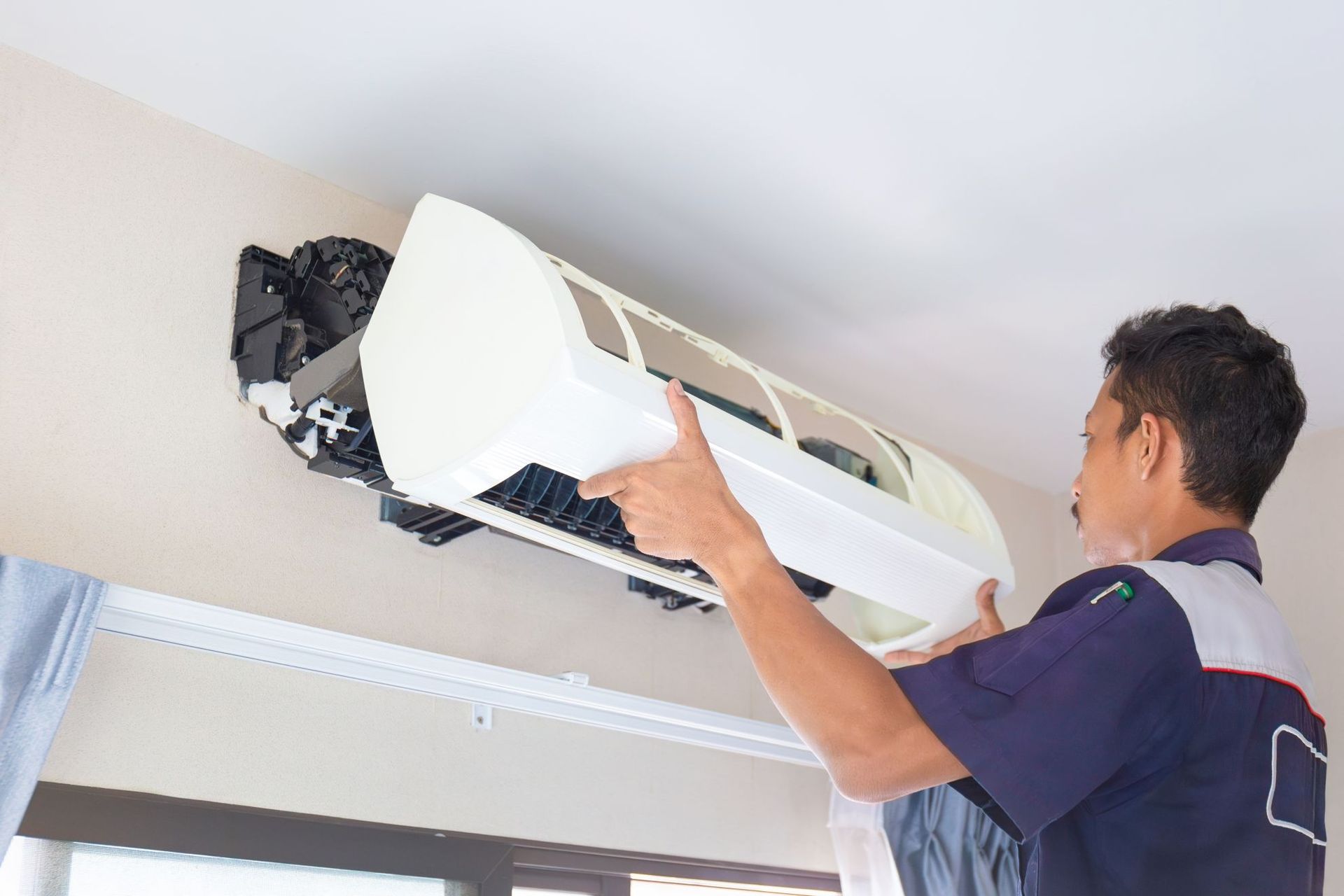 A technician in a uniform repairs a white split-system air conditioner mounted on an indoor wall.