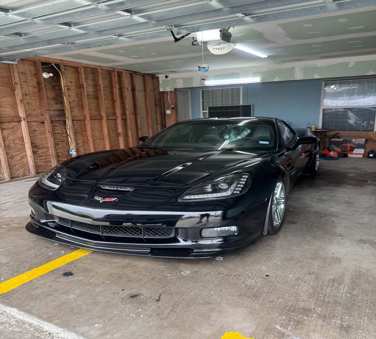 A black Chevrolet Corvette parked inside a residential garage with unfinished wooden wall framing.
