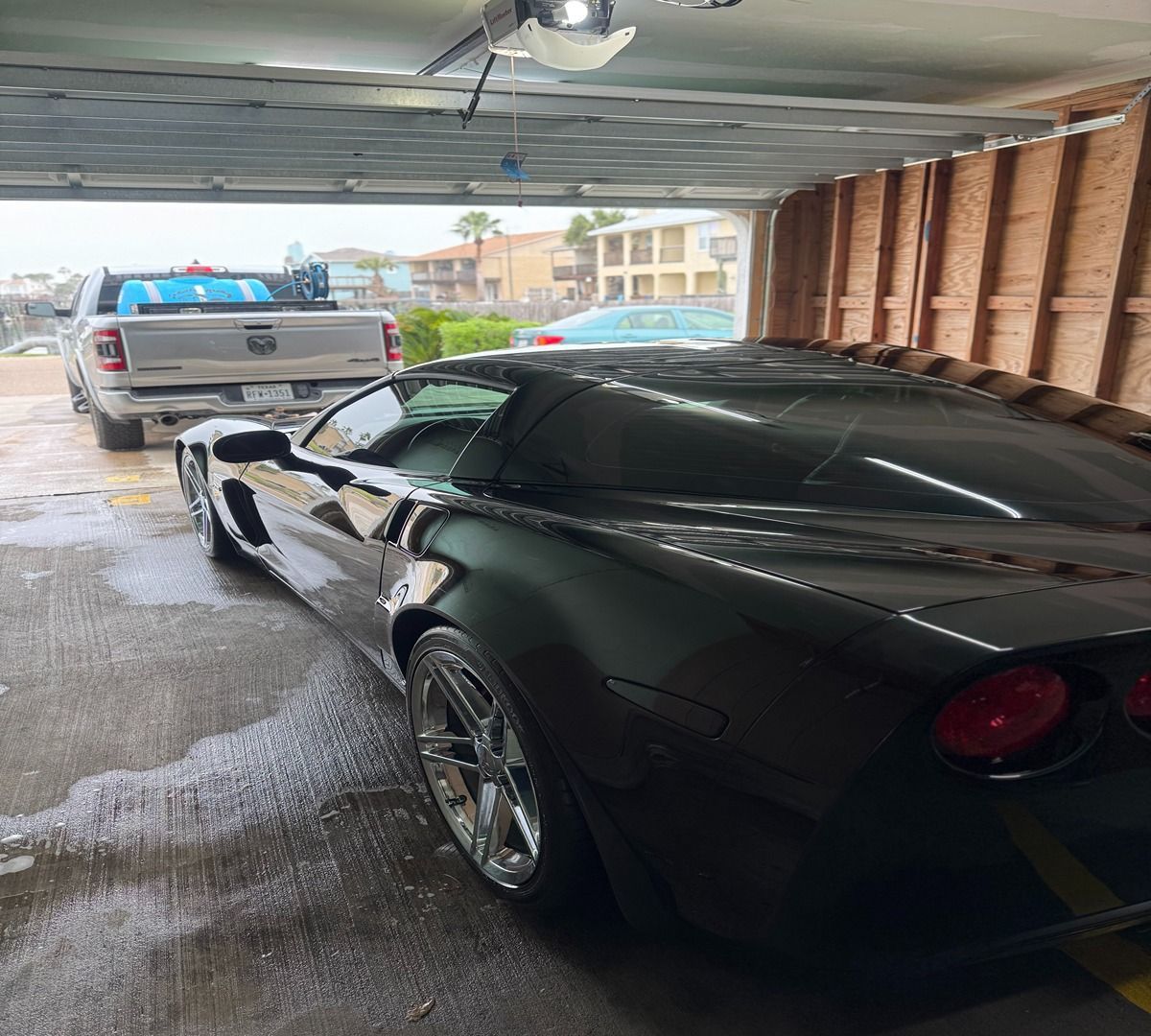A shiny black Corvette parked in a garage, with a light-colored pickup truck visible in the background outside.