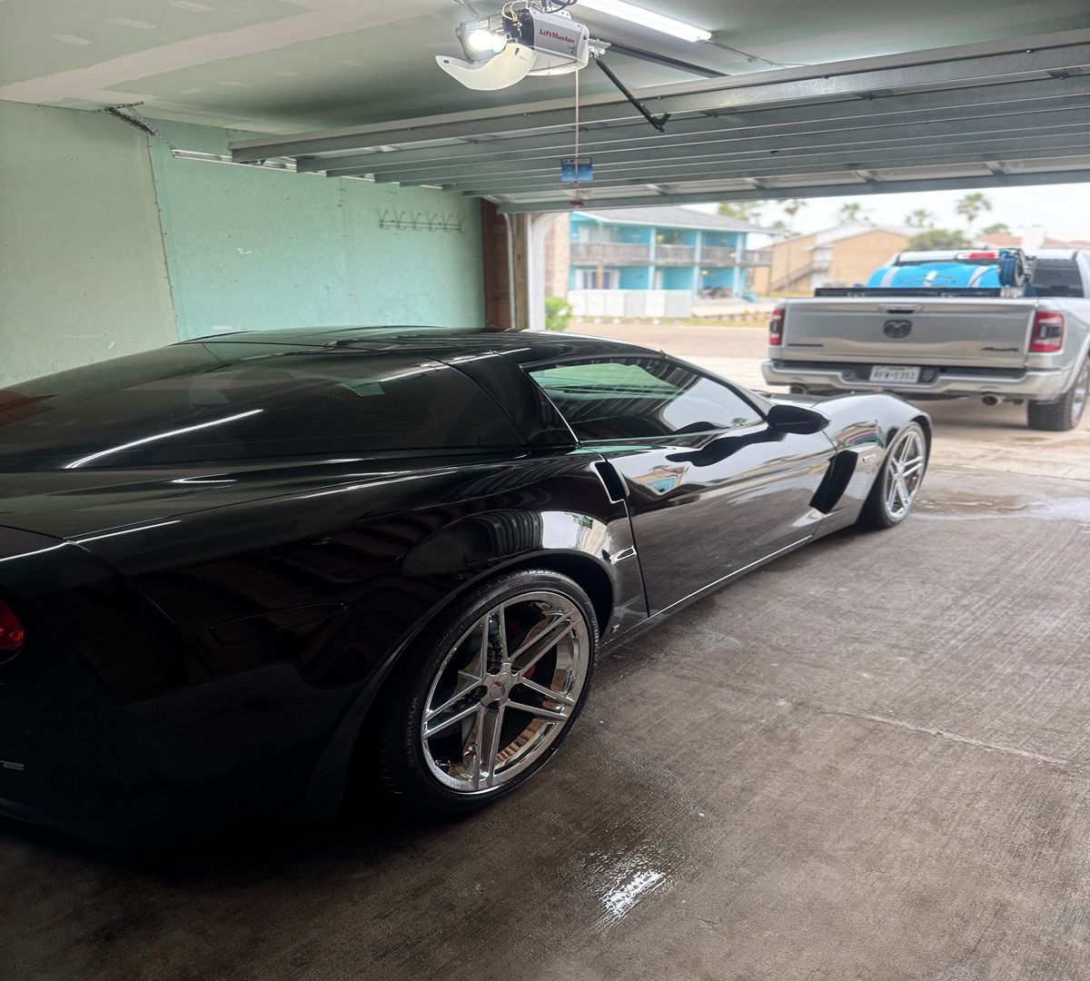 A black sports car parked inside a garage, with a white pickup truck visible outside in the driveway.