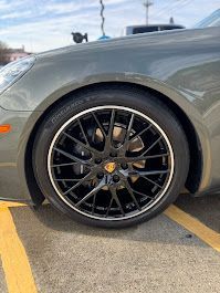 A close-up view of a matte grey Porsche's front wheel, featuring a black multi-spoke rim with a central Porsche emblem.