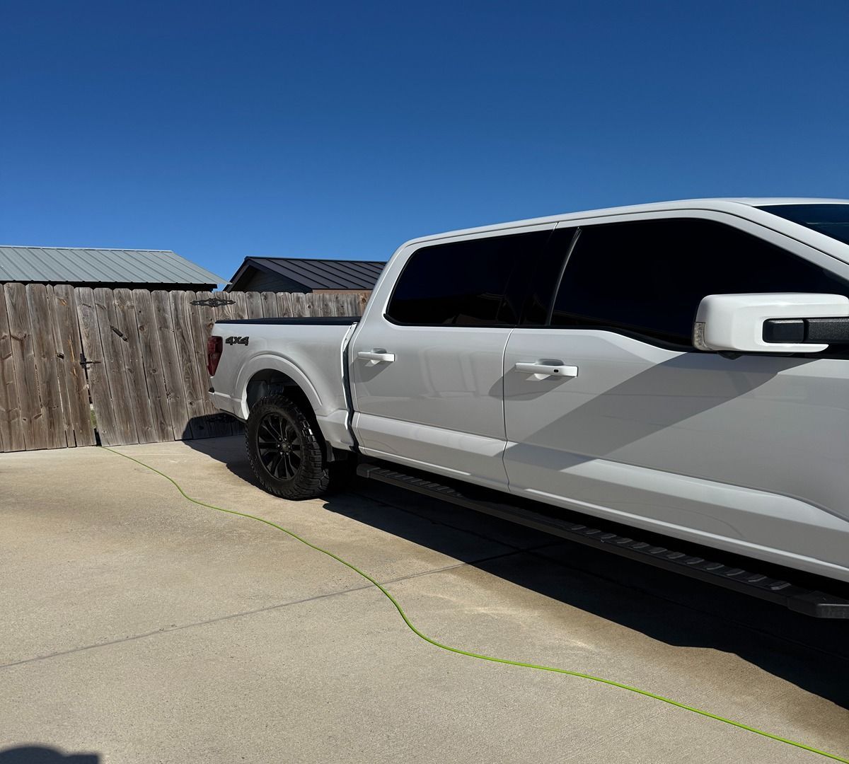 A side view of a white Ford F-150 pickup truck parked on a concrete driveway next to a wooden fence.