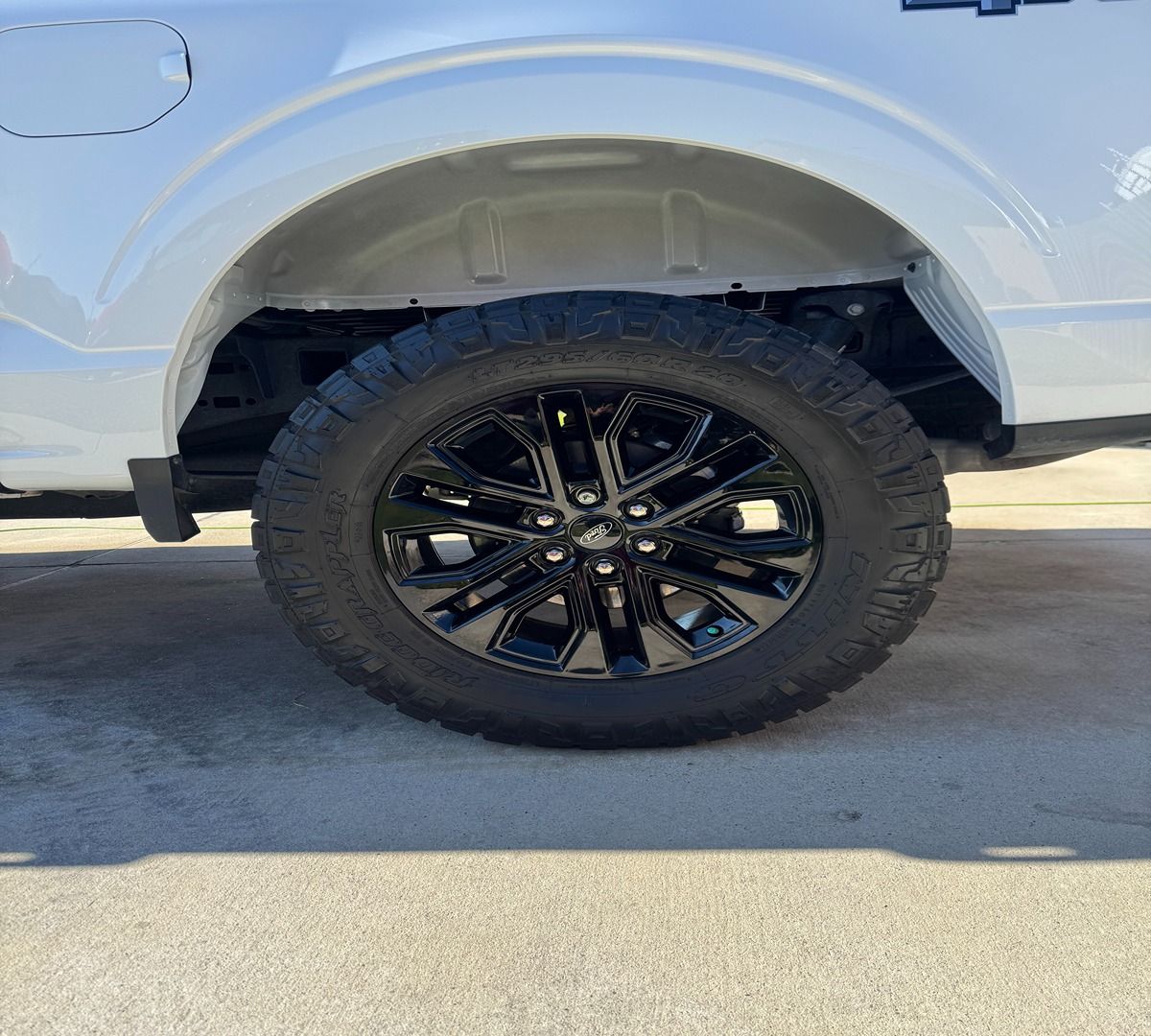 A white pickup truck’s rear wheel with a black, multi-spoke alloy rim and an all-terrain tire on a concrete surface.