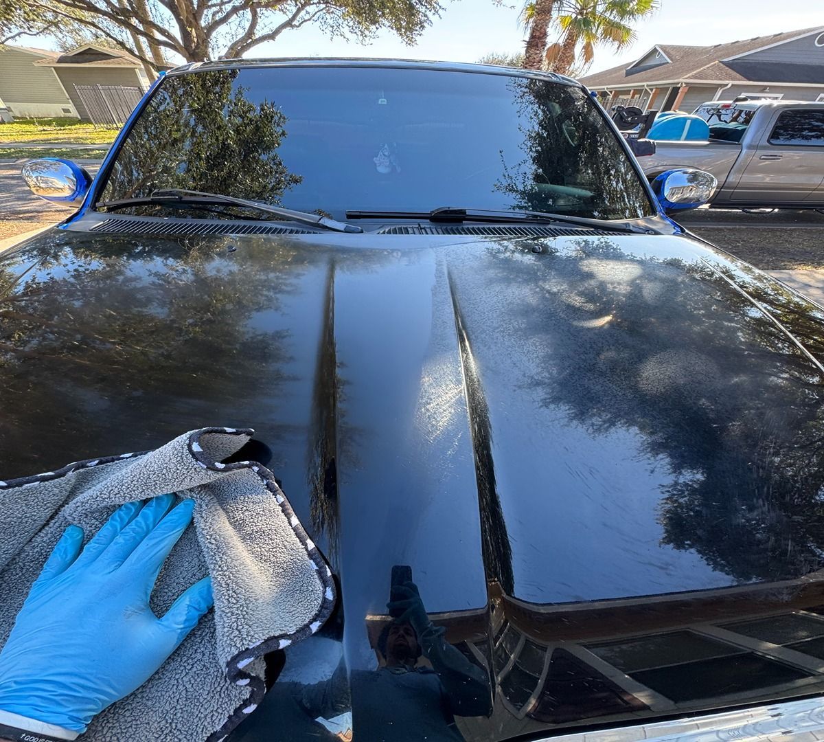 A gloved hand uses a grey microfiber cloth to buff the glossy black hood of a parked vehicle.