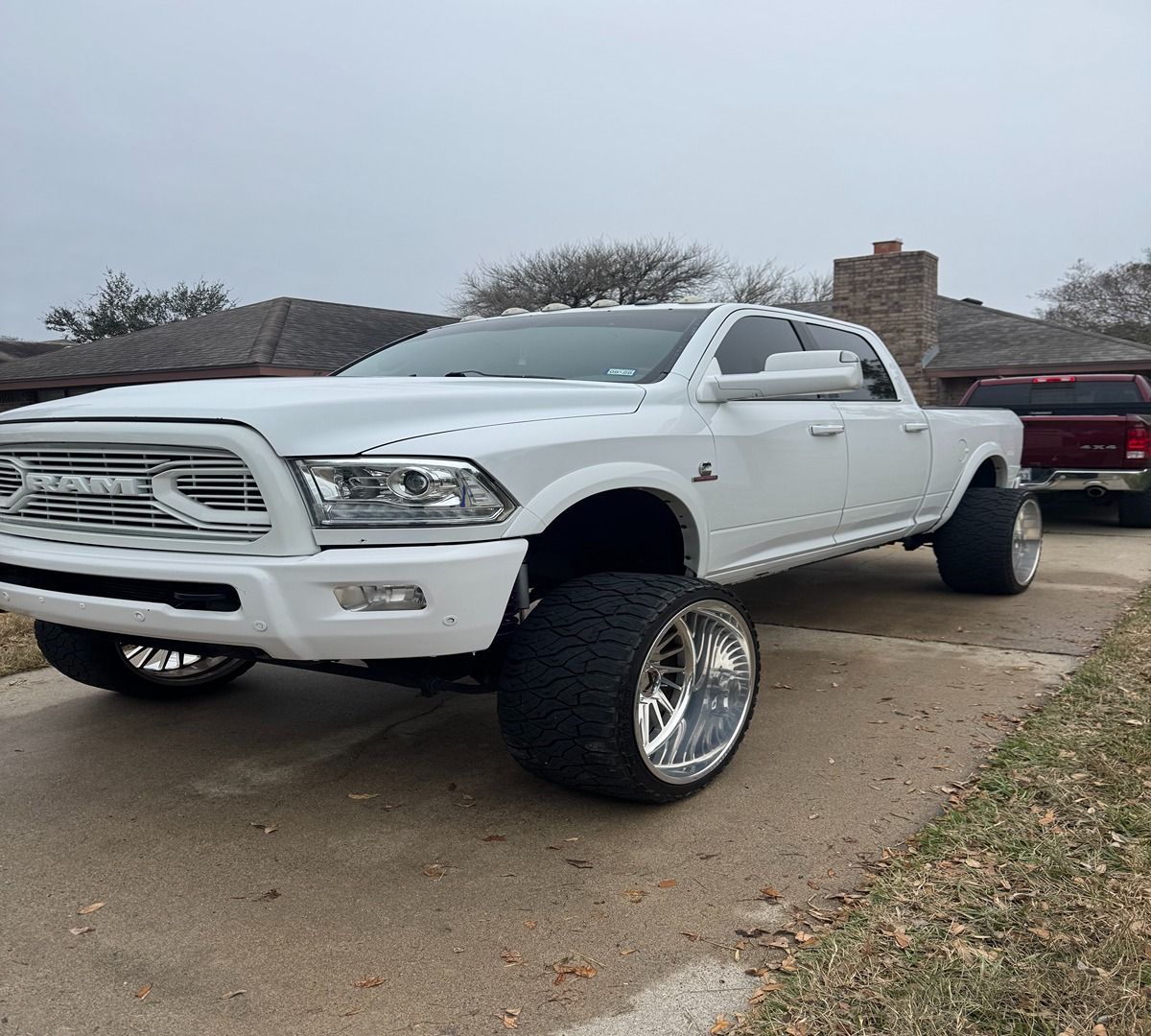 A white Ram pickup truck with a lifted suspension and wide, aftermarket wheels parked on a suburban concrete driveway.