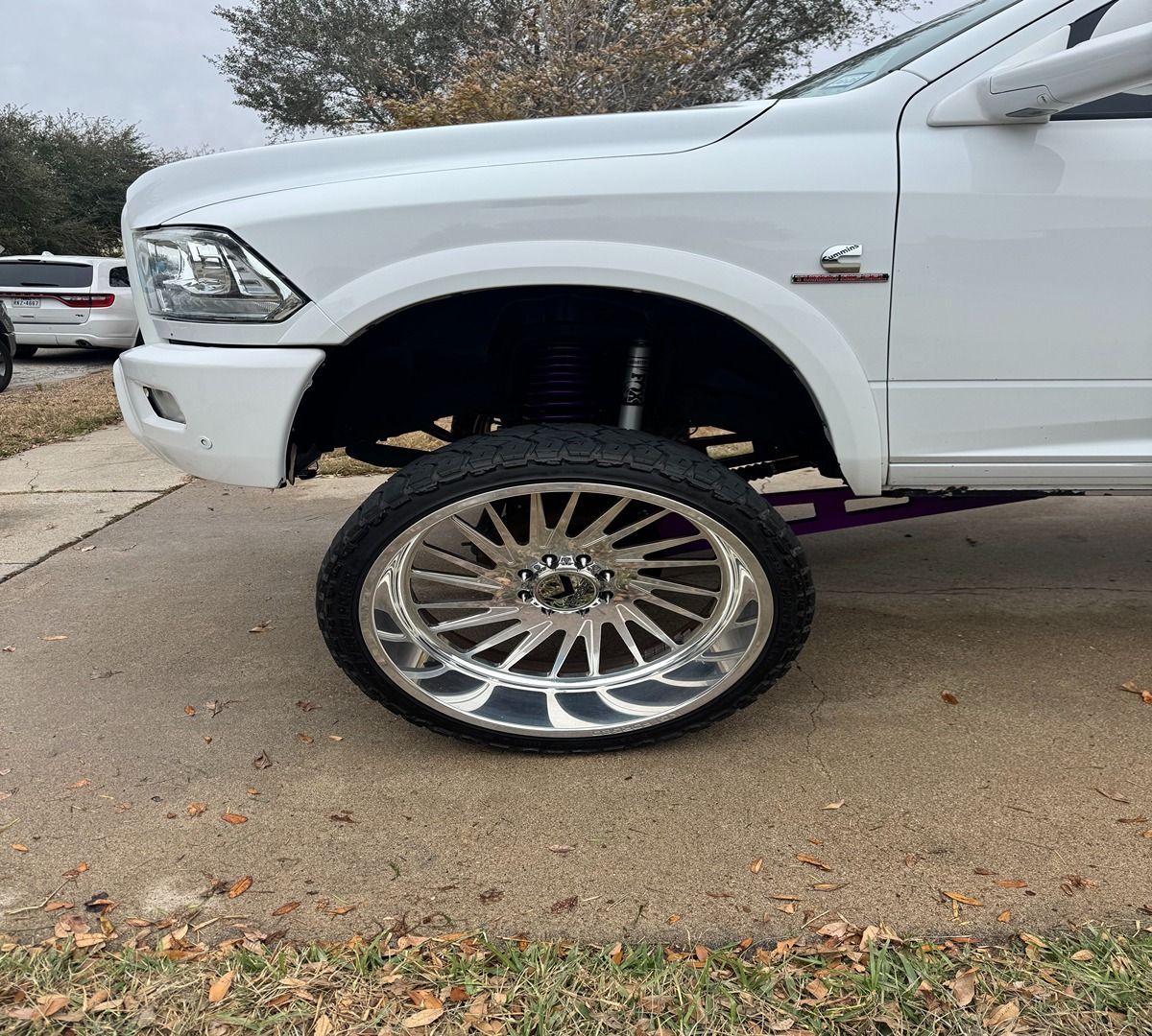 A white lifted pickup truck on a concrete driveway, featuring large, polished chrome multi-spoke wheels.