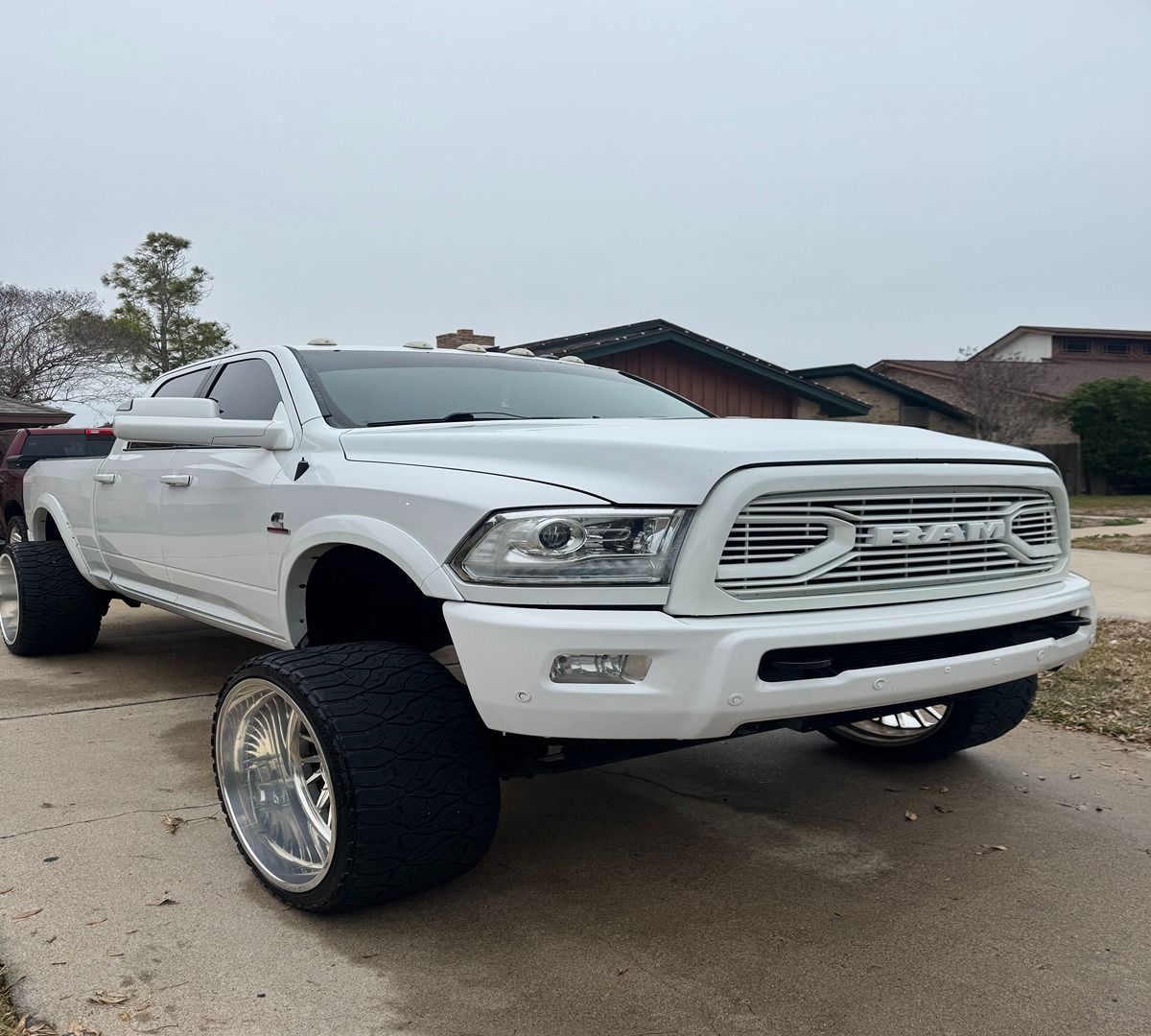 A modified white Ram pickup truck parked on a residential driveway, featuring oversized tires and a chrome grille.